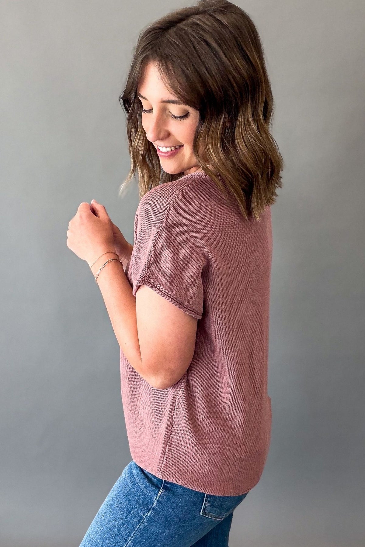 A woman with shoulder-length brown hair is shown from the back wearing a mauve short-sleeve knit top and blue jeans against a plain gray studio background.