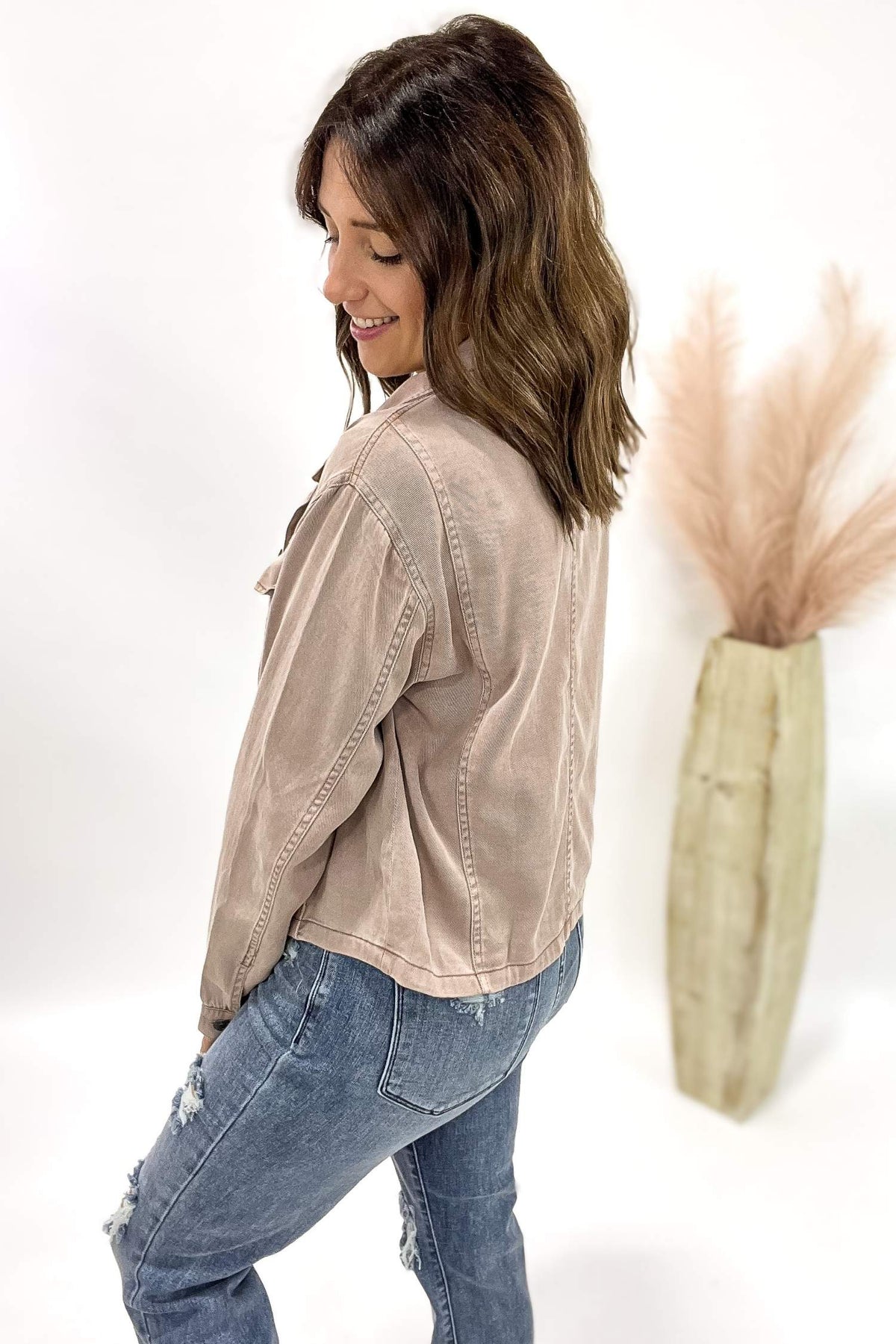Woman wearing a dusty mauve jacket and blue jeans standing next to a vase with dried plants on a white background