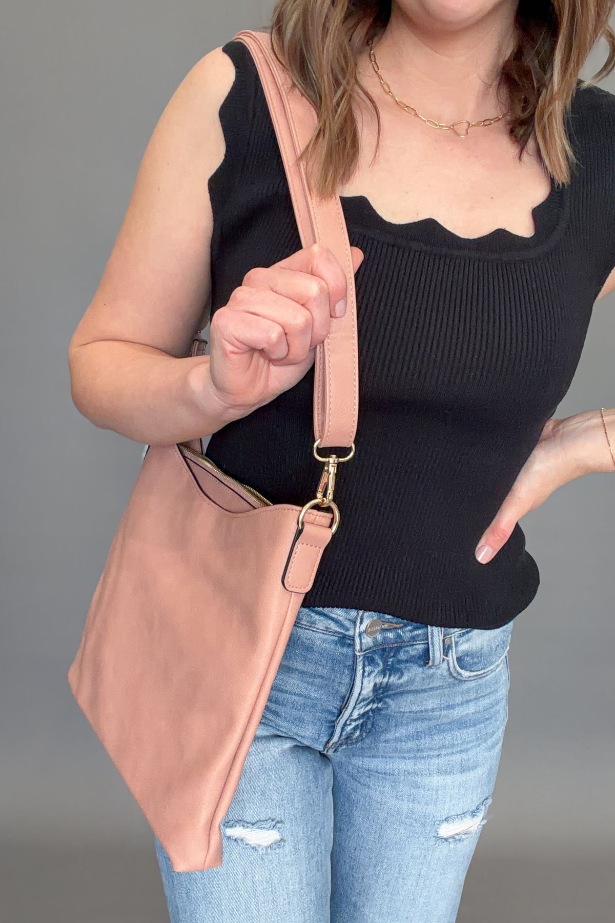 Person wearing a black tank top and blue jeans, holding a mauve shoulder bag against a gray background