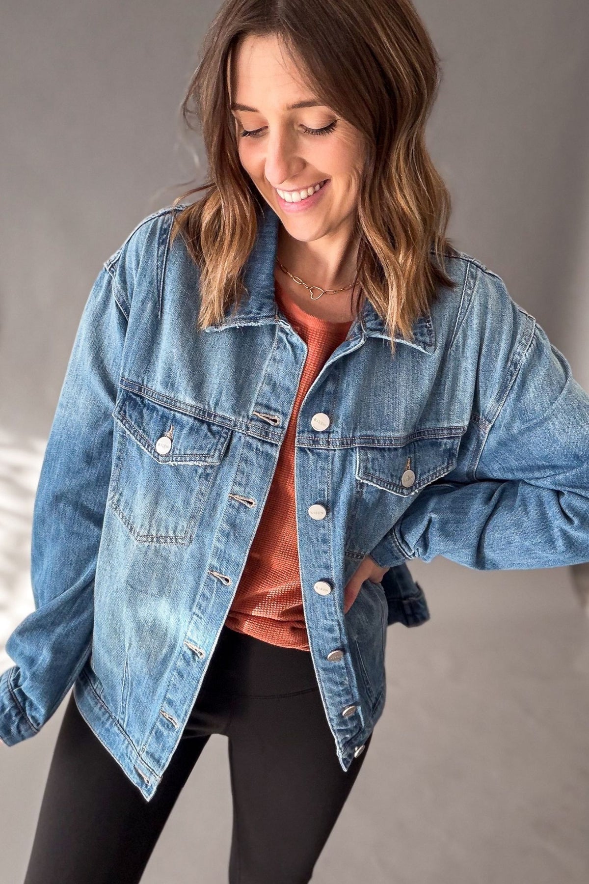 A woman wearing a medium blue denim jacket over a rust-orange top and black leggings, smiling and looking down against a plain gray studio background.
