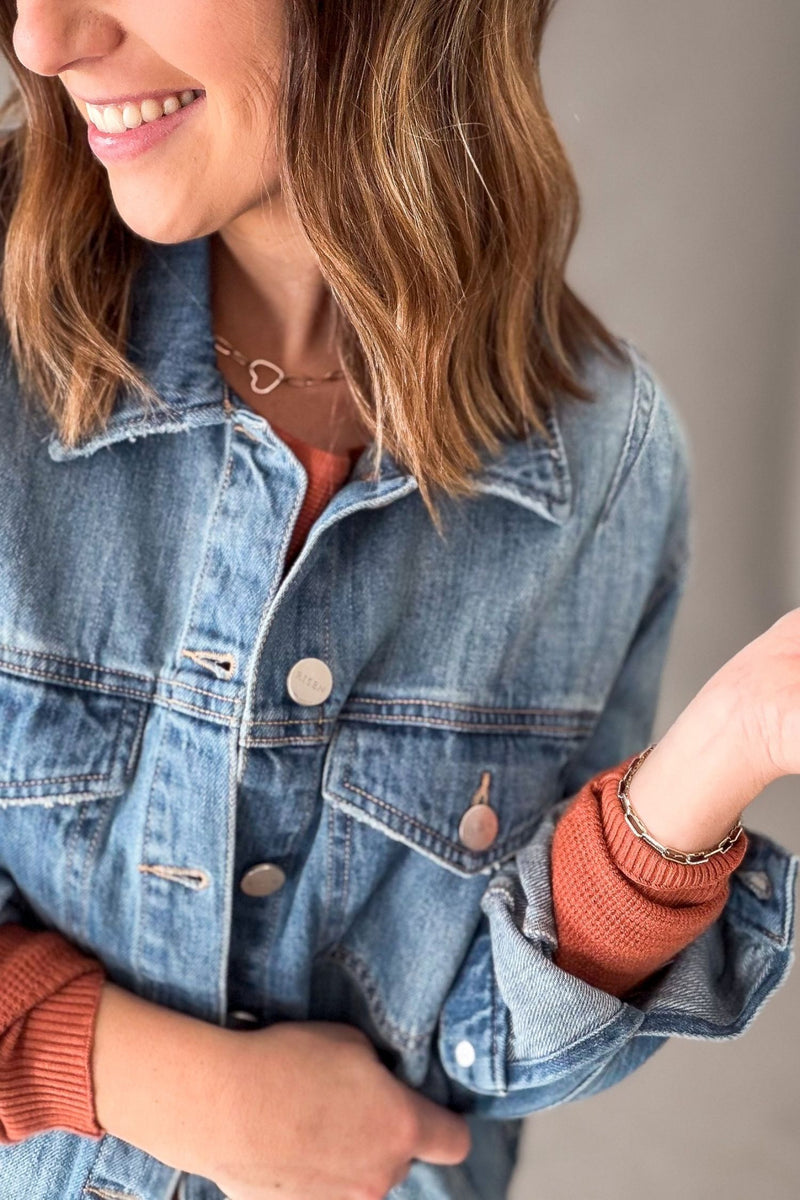 A close-up of a woman wearing a medium blue denim jacket over a rust-orange ribbed sweater, with her hair, smile, and hands visible against a soft neutral background.