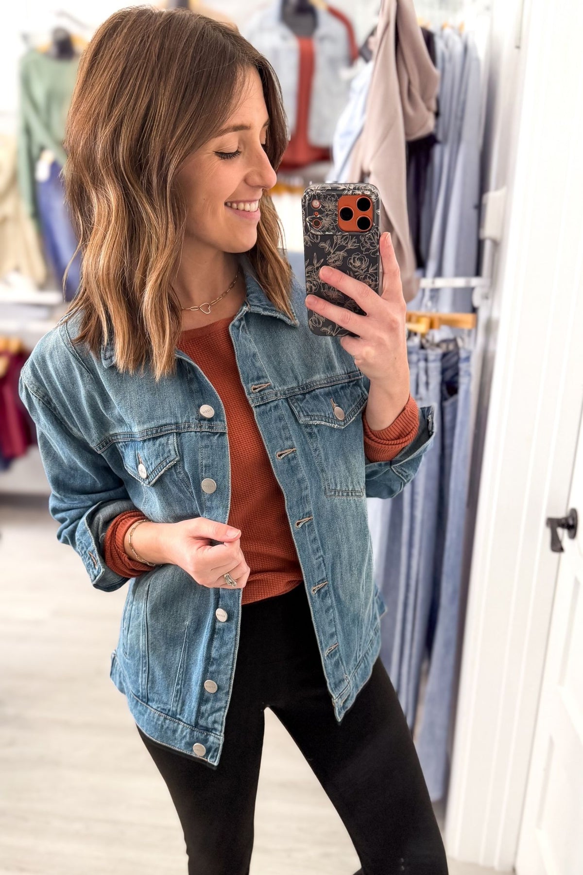 A woman takes a mirror selfie in a clothing store, wearing a medium-wash blue denim jacket over a rust-orange long-sleeve top and black pants.