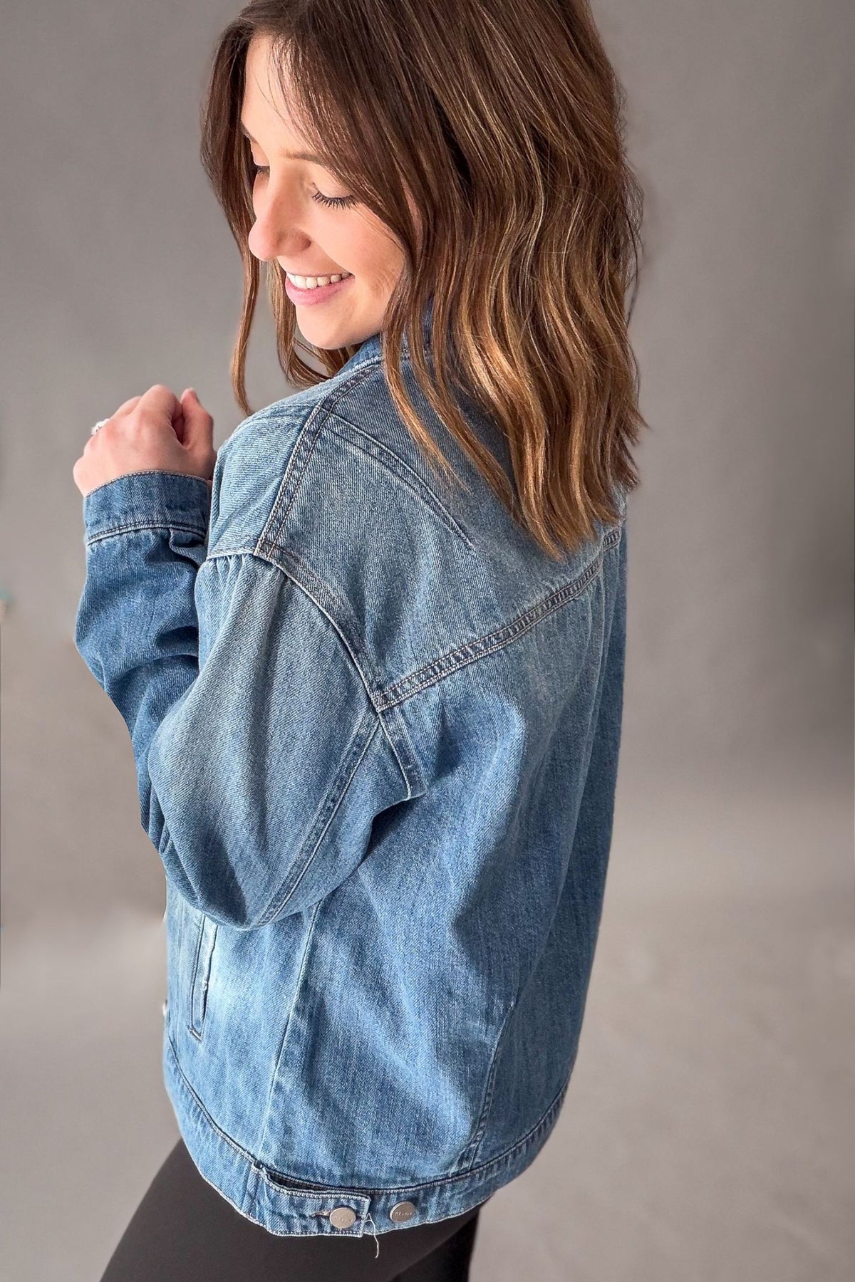 A woman wearing a medium blue denim jacket, shown from a side-back angle against a neutral gray studio background.