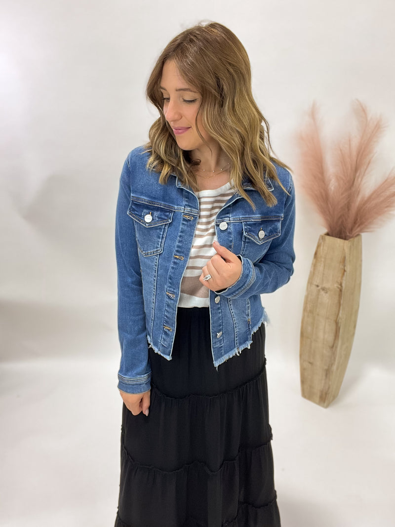 Woman wearing a denim jacket over a striped shirt with a black skirt, standing against a white wall.