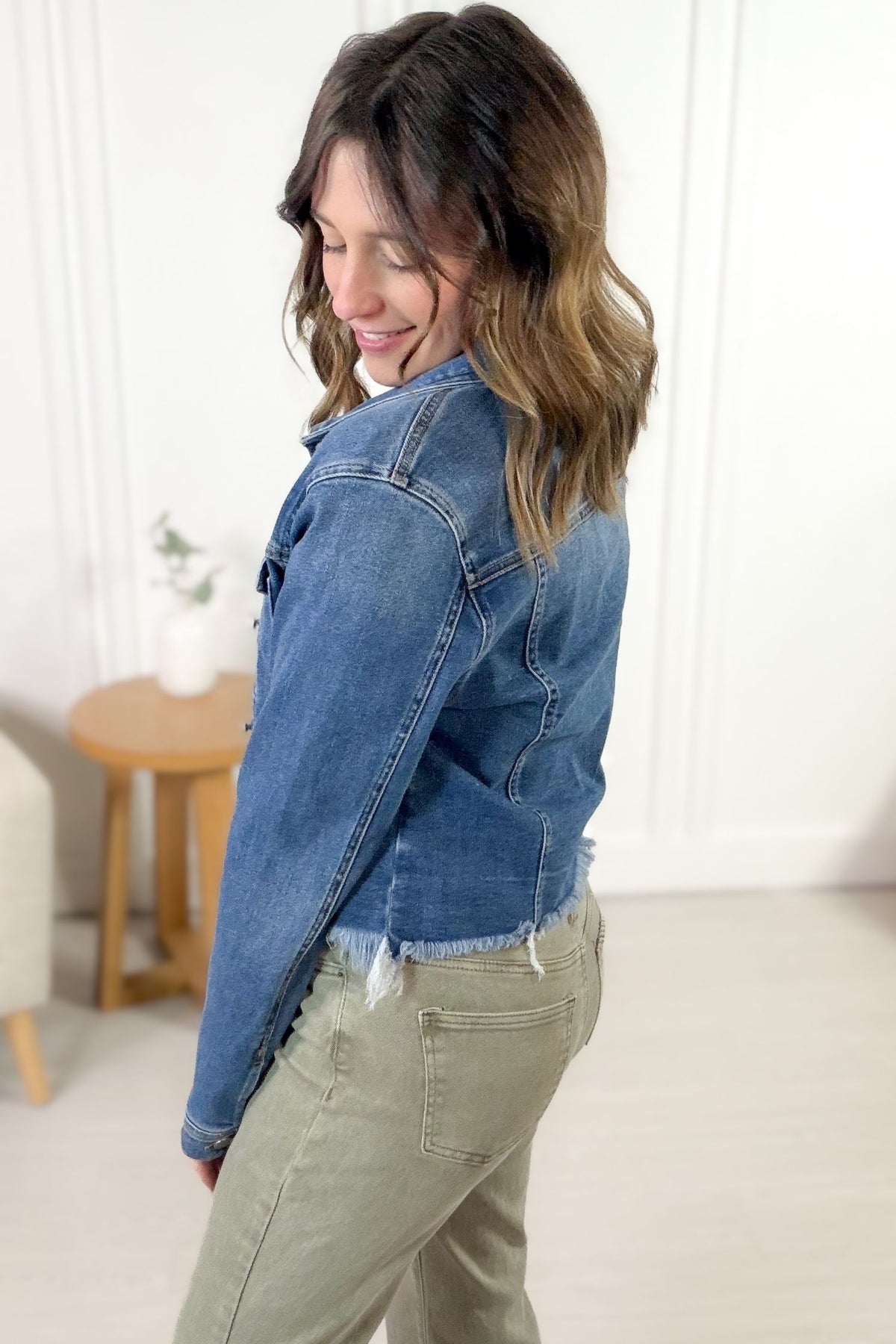 Woman wearing a blue denim jacket and green pants in a room with a white wall and wooden table.
