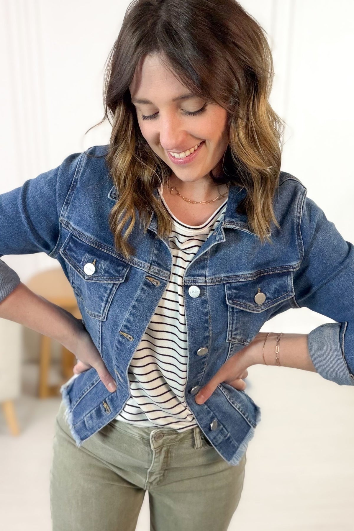 Woman wearing a denim jacket, striped shirt, and green pants indoors.
