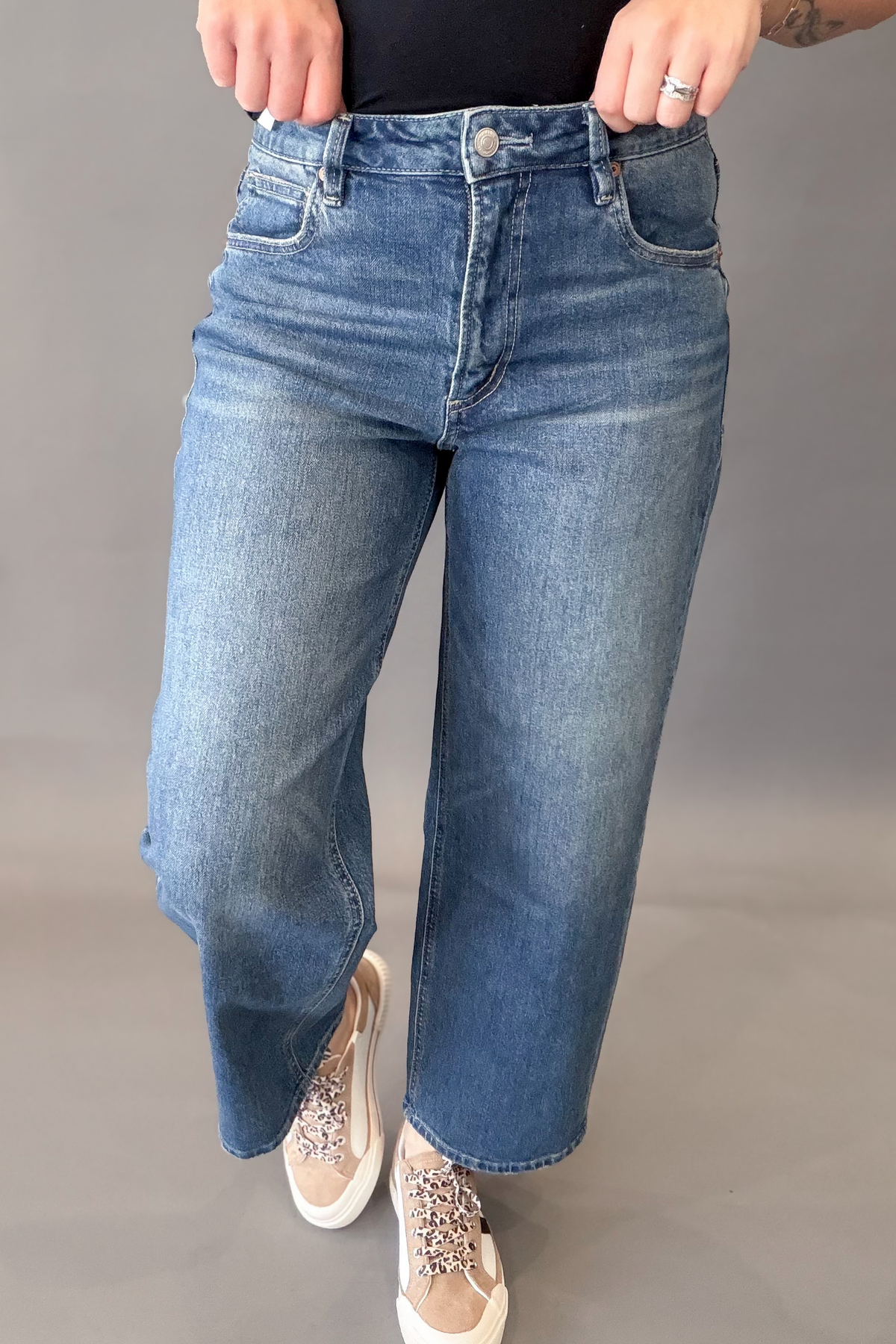 A person wearing medium-wash blue denim jeans with a straight-to-wide leg fit, paired with beige embellished sneakers, shown from the waist down against a neutral gray studio background.