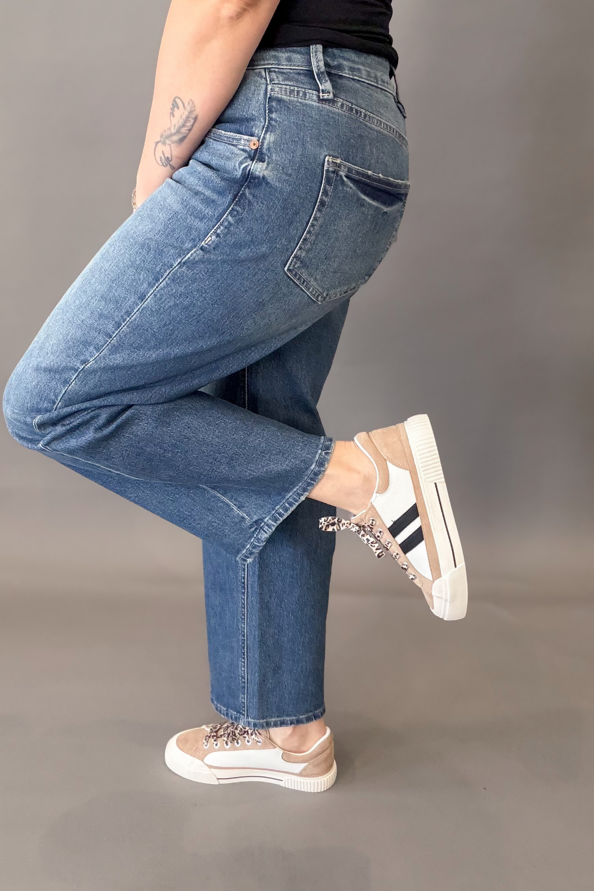 A person wearing blue straight-leg jeans and beige-and-white sneakers with black stripes, shown from the waist down against a plain gray studio background.