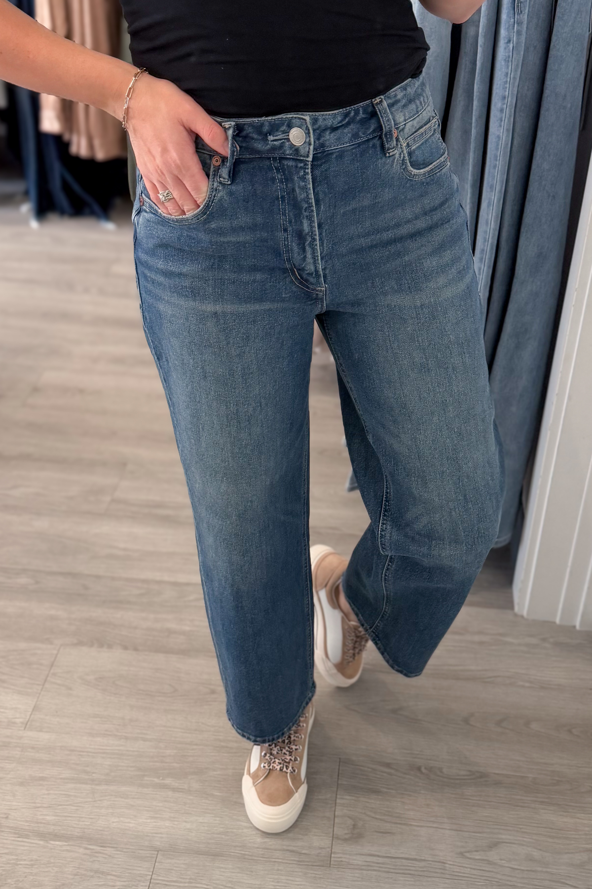 A person wearing medium-wash blue jeans with a straight-to-wide leg fit, paired with a black top and beige sneakers, standing in a clothing store.