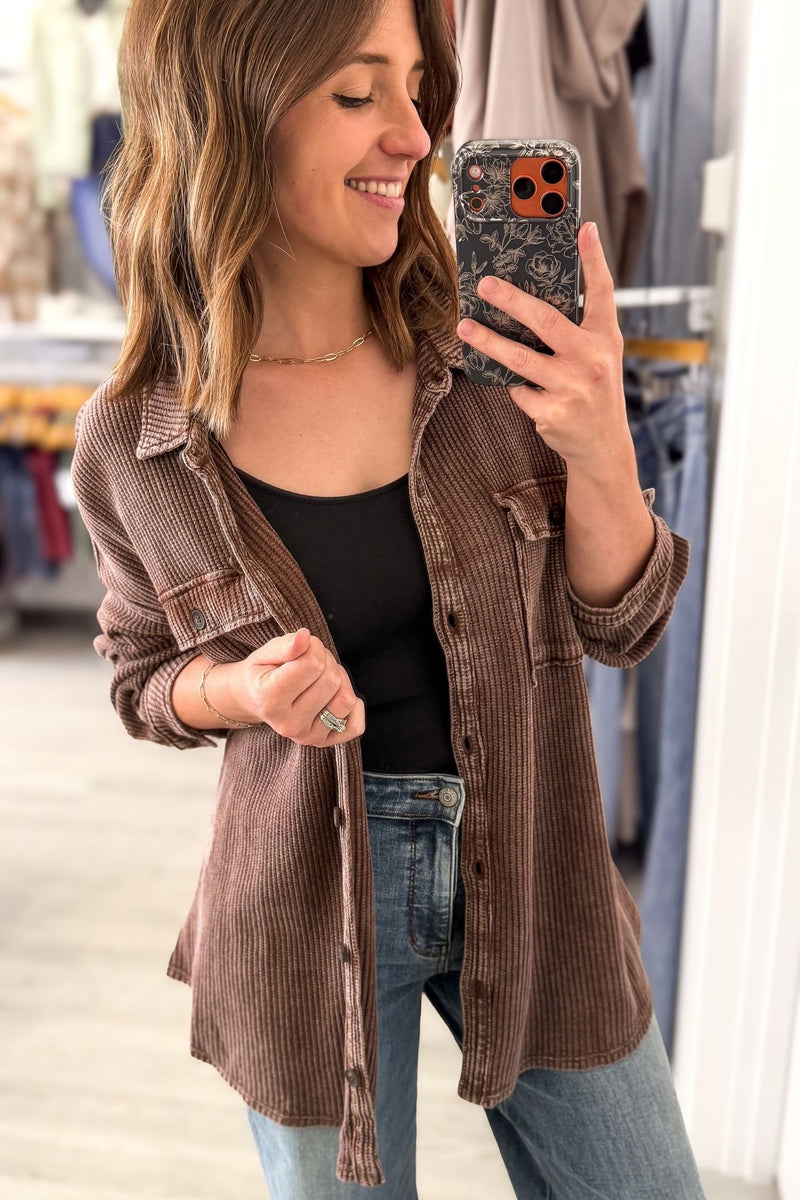 A woman takes a mirror selfie in a clothing store while wearing a brown ribbed button-up shirt over a black tank top and blue jeans.