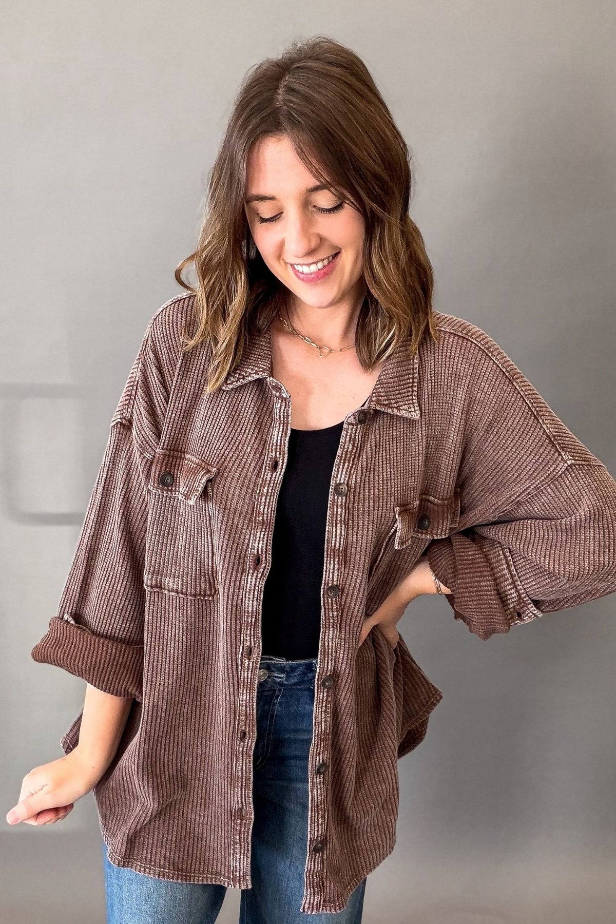 A woman wearing an oversized brown ribbed button-up shirt over a black top and blue jeans, standing against a plain gray wall.