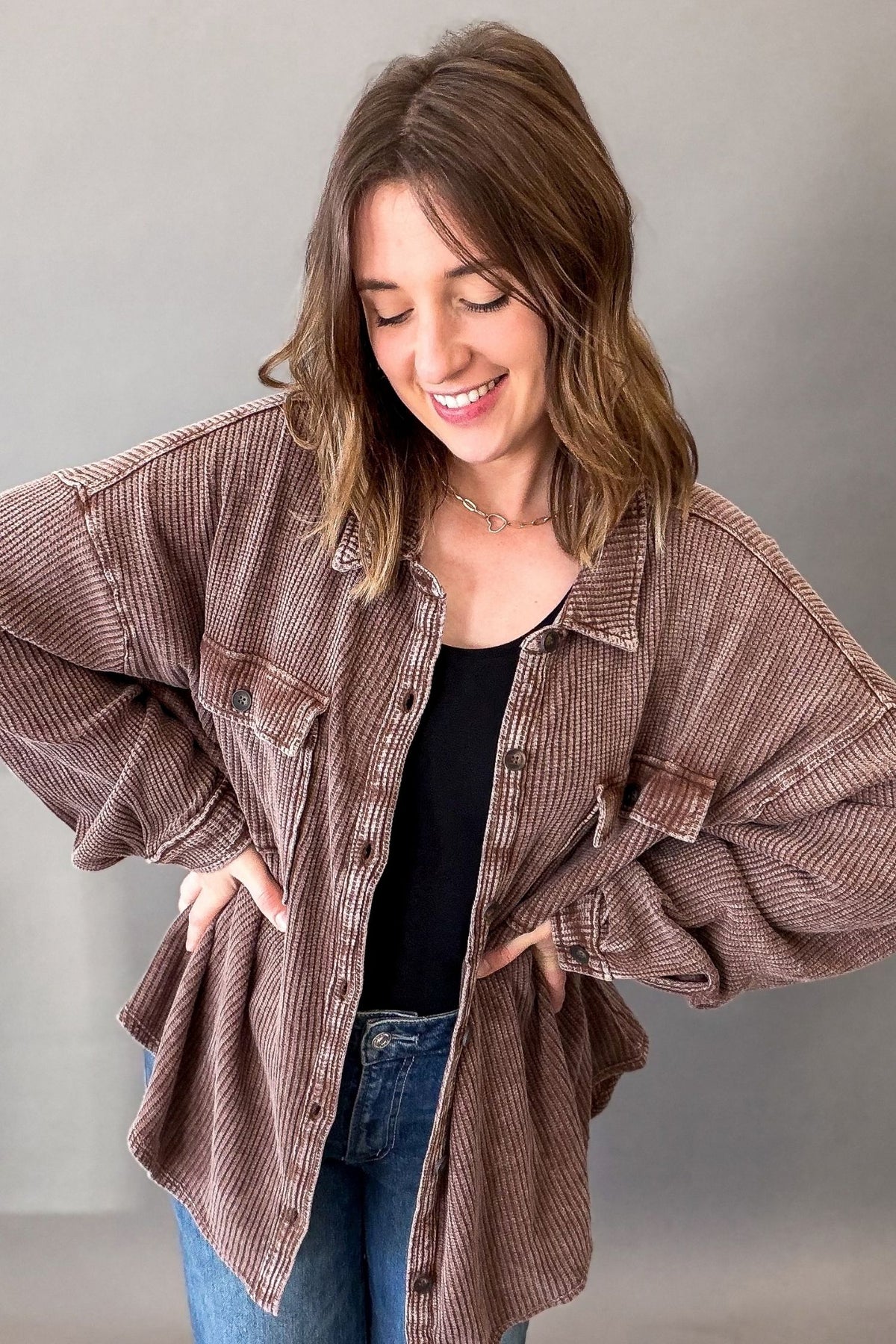 A woman wearing an oversized brown ribbed button-up shirt over a black top and blue jeans, standing against a plain gray background.
