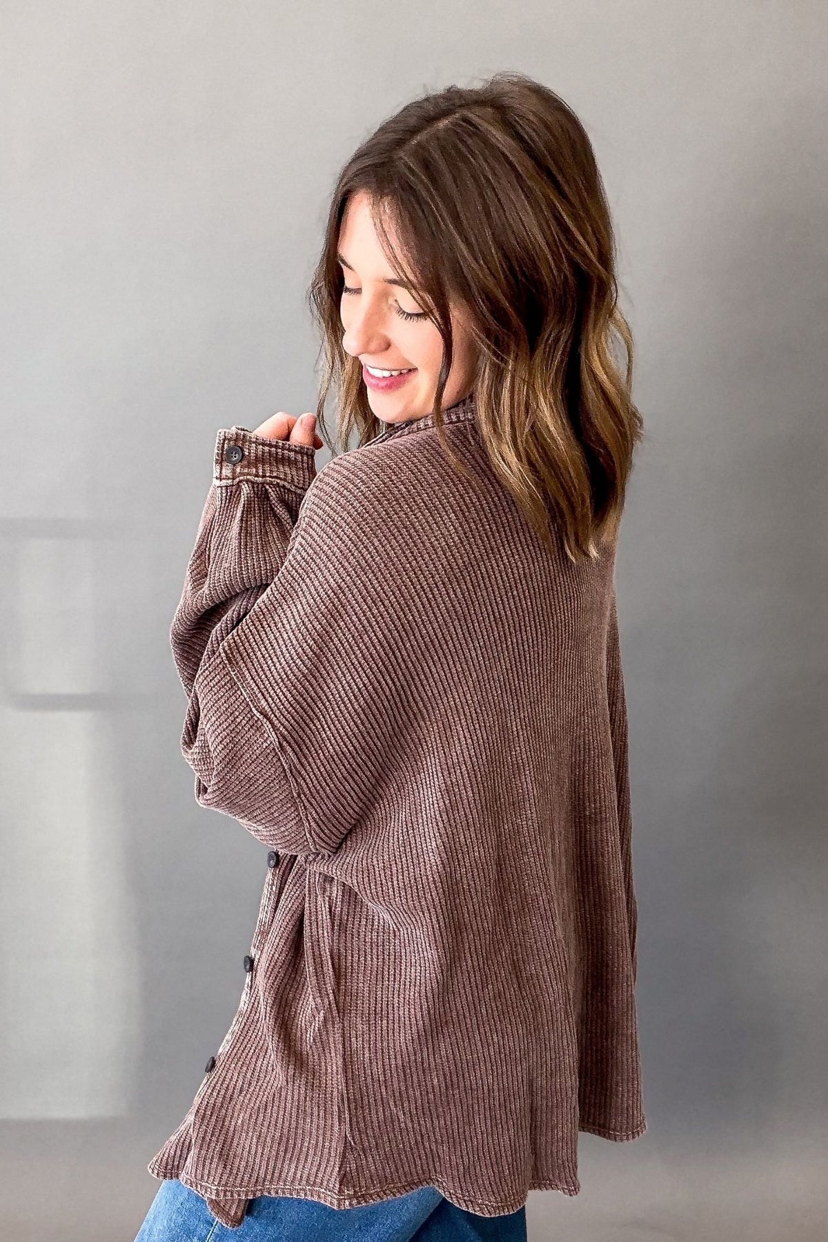 A woman wearing a brown ribbed button-up shirt with blue jeans, photographed from the back against a neutral gray background.