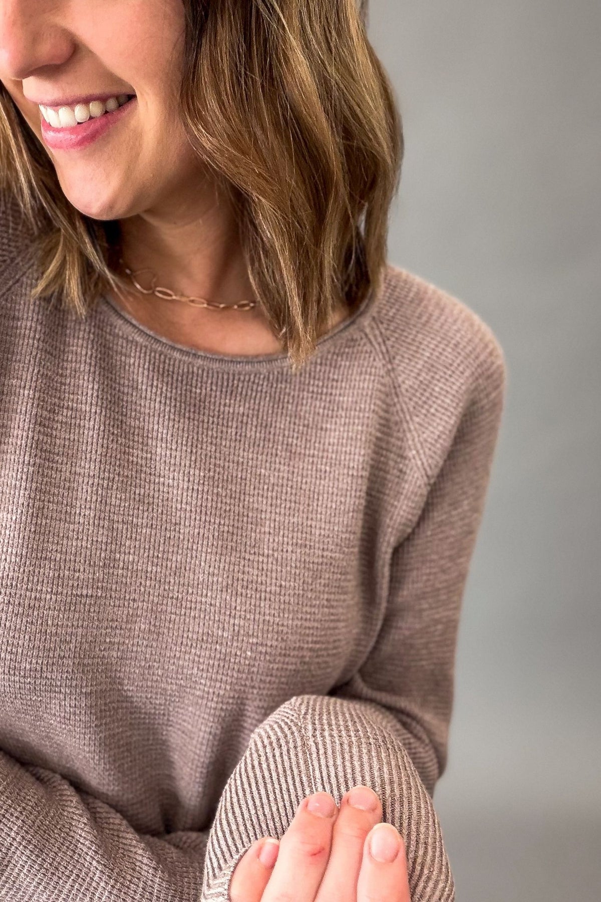 A close-up of a woman wearing a light mocha mini waffle knit crewneck sweater, smiling and showing her hands and hair against a neutral gray background.