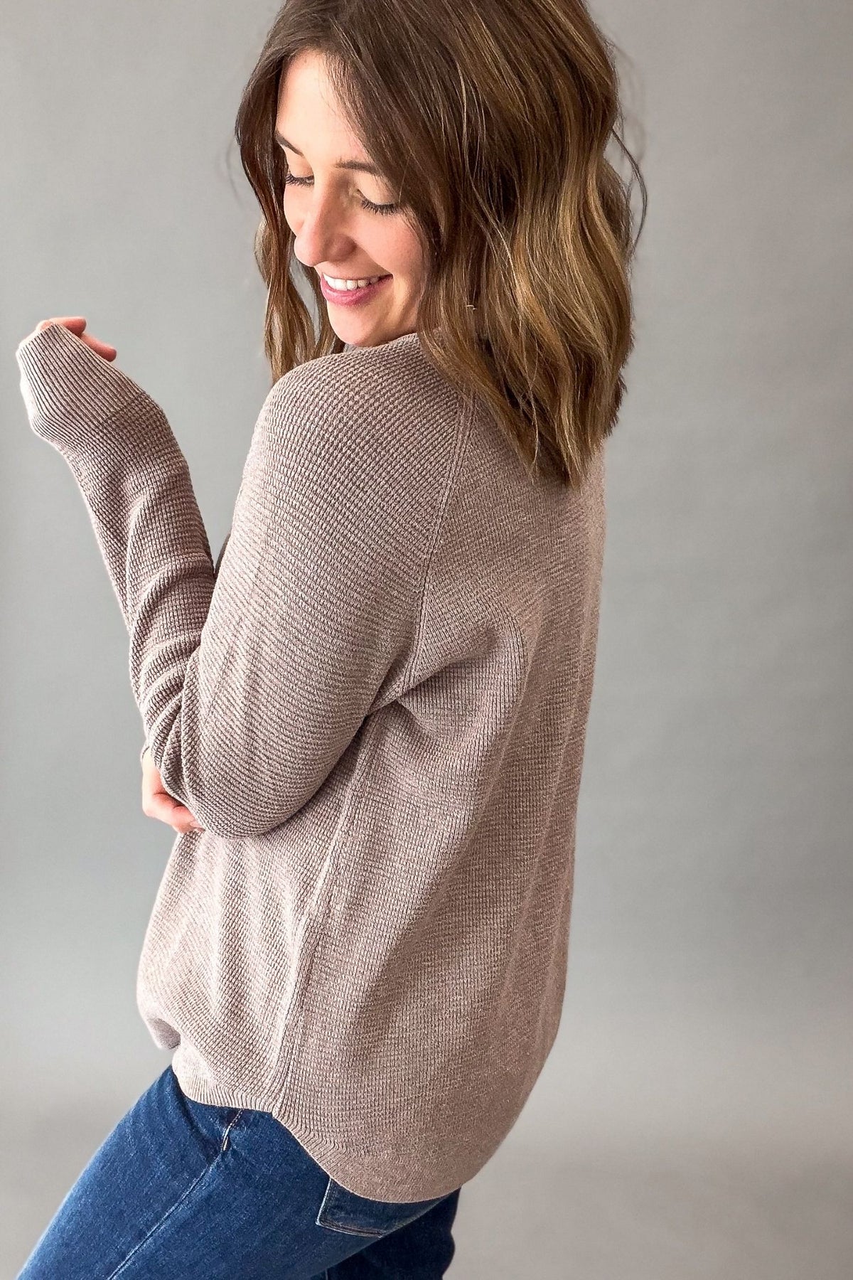 A woman wearing a mini waffle light mocha knit long-sleeve sweater,  paired with blue jeans, photographed against a neutral gray studio background.