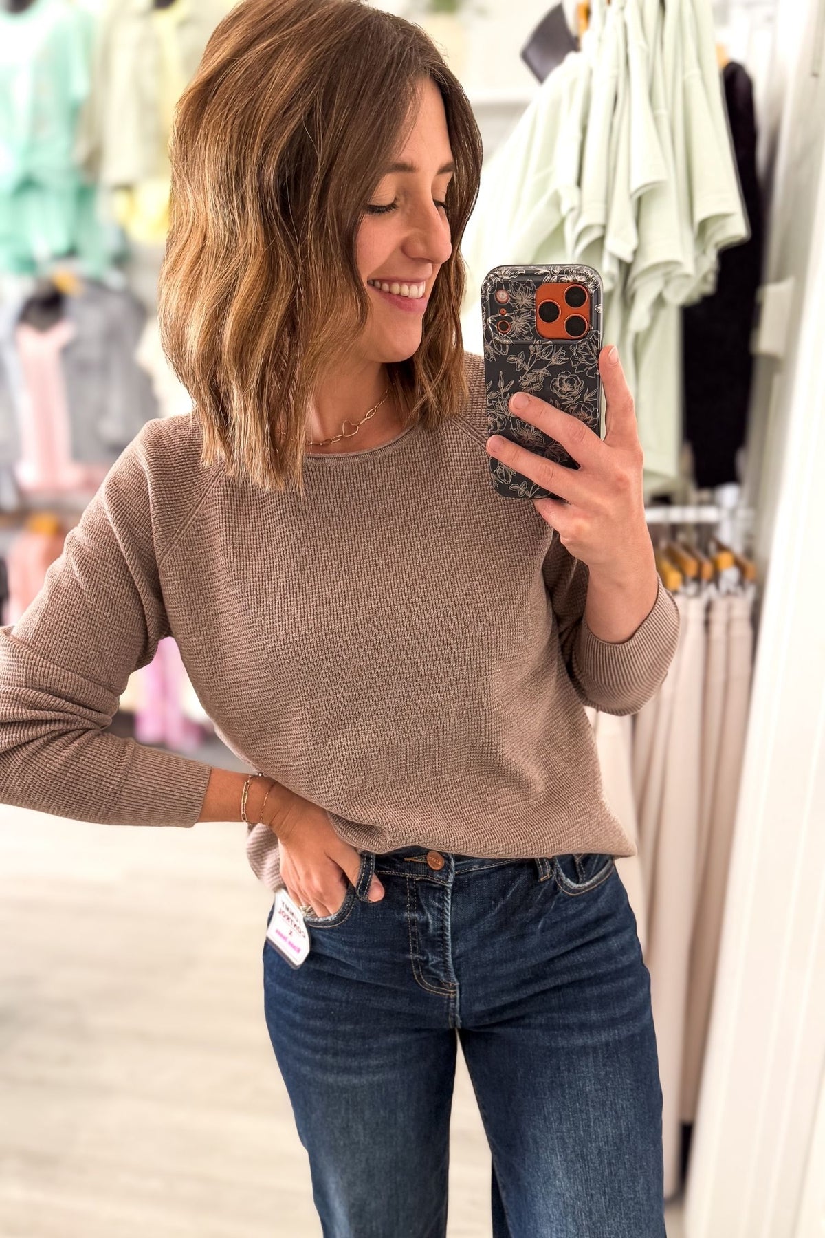 A woman takes a mirror selfie in a clothing store, wearing a mini waffle knit long-sleeve sweater and dark wash blue jeans.