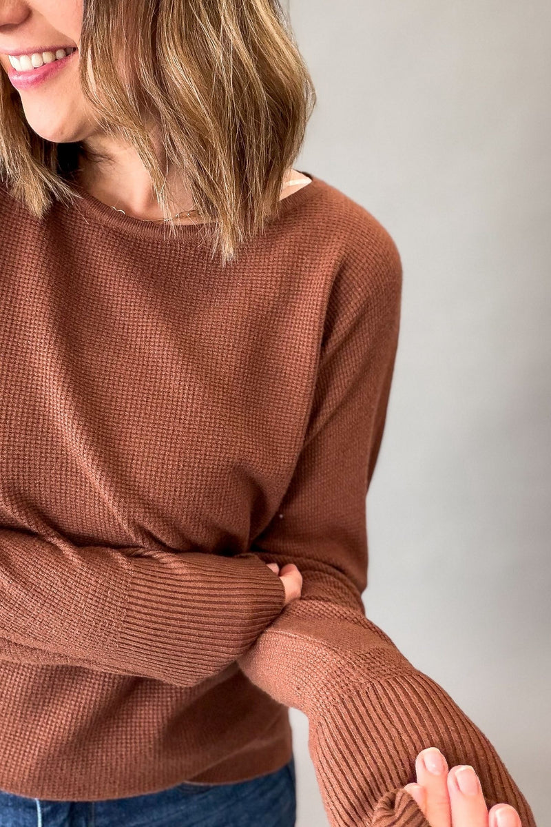A close-up of a woman wearing a dark copper mini waffle knit sweater, showing her smiling, hair, and hands adjusting the sleeve.