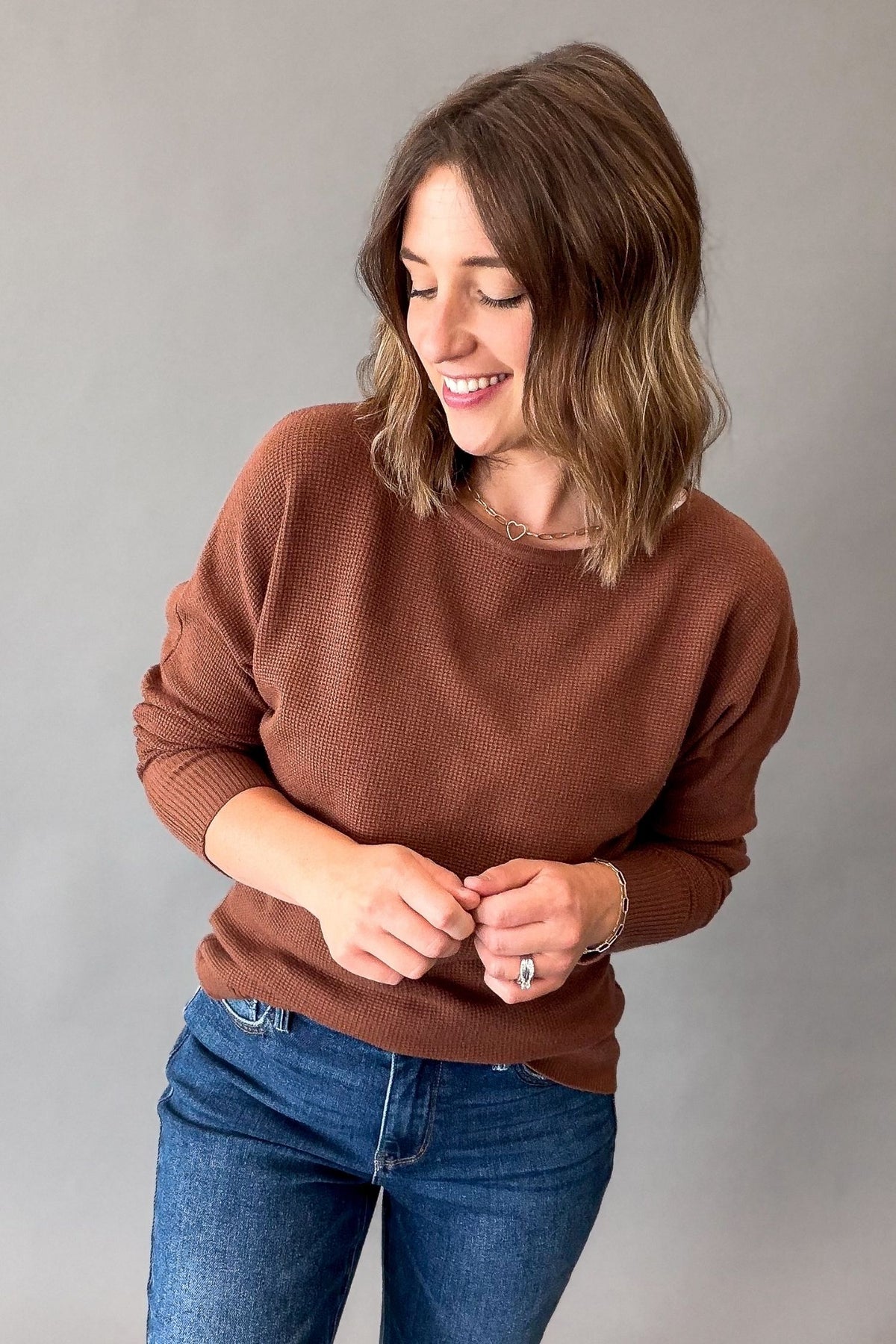 A woman wearing a dark copper ribbed knit crewneck sweater and blue jeans, standing against a plain gray studio background.