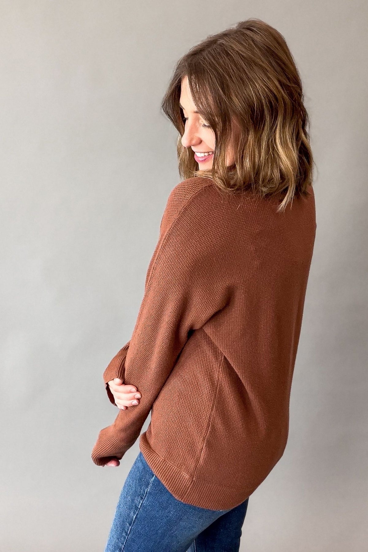 A woman wearing a dark copper mini waffle knit sweater and blue jeans, photographed from the back against a plain gray studio background.