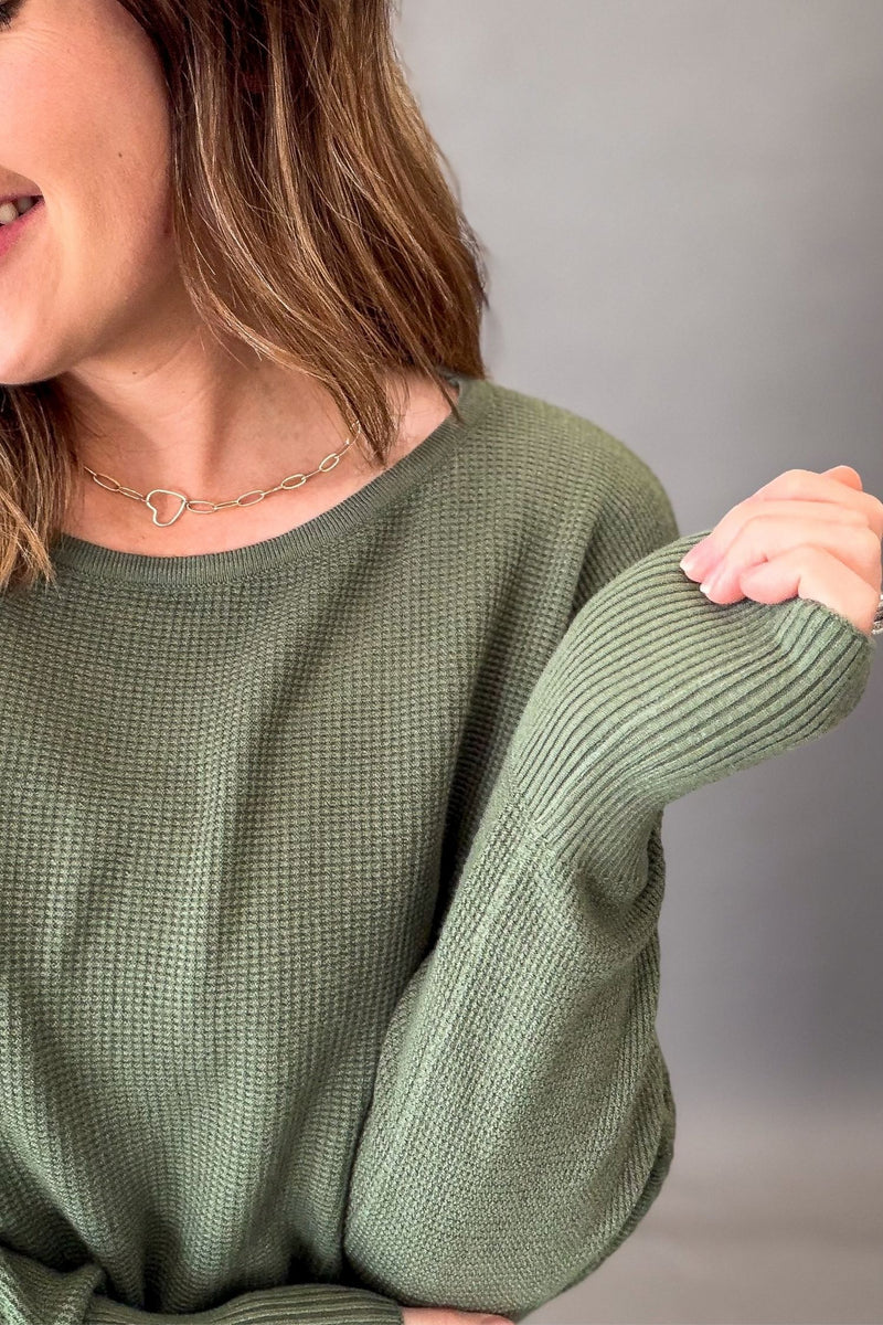 A close-up of a woman wearing an olive green waffle-knit sweater, showing the texture of the fabric and the ribbed cuffs, with a neutral gray background.