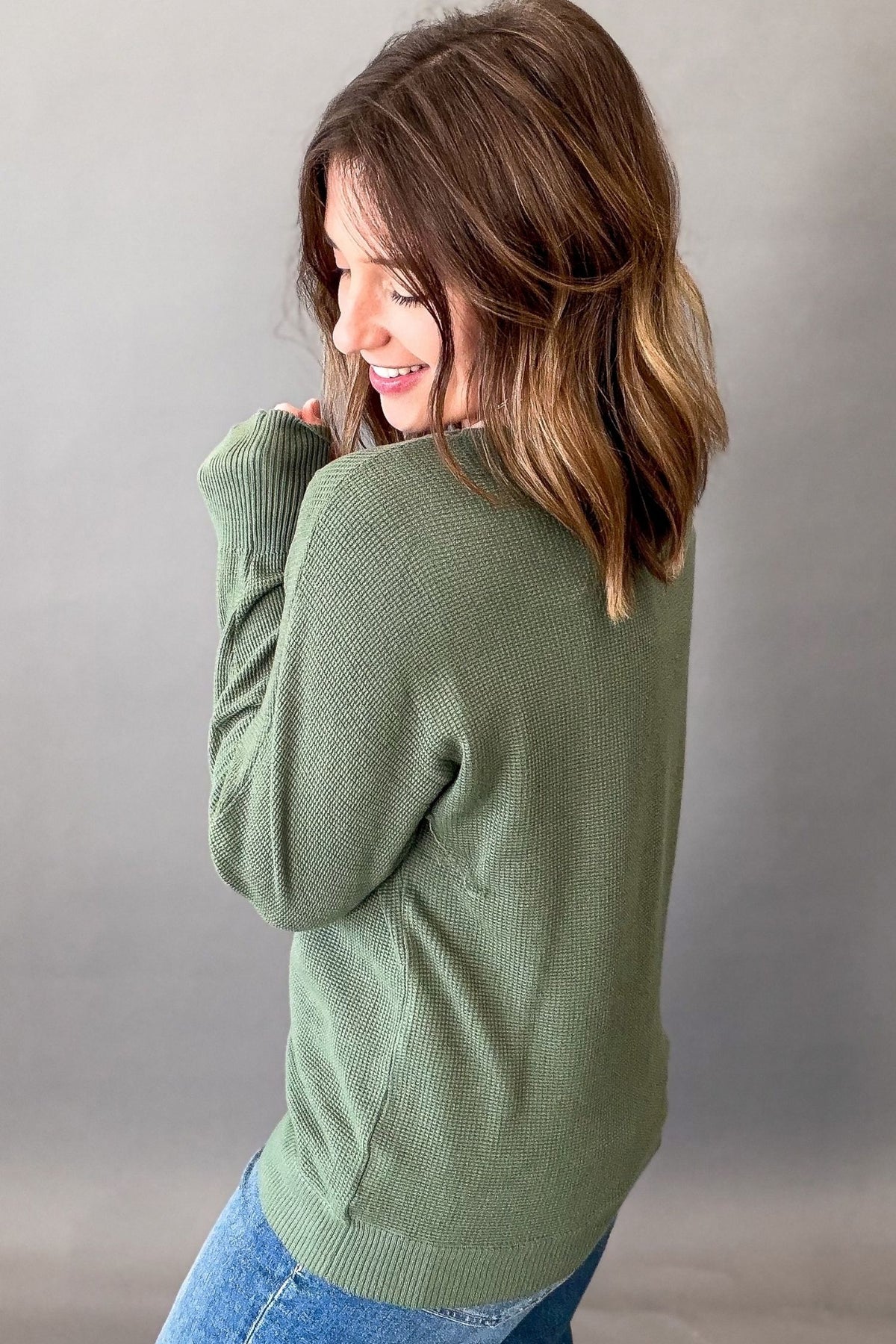 A woman with shoulder-length brown hair is shown from the back wearing an olive green ribbed knit sweater and blue jeans, posing against a neutral gray studio backdrop.