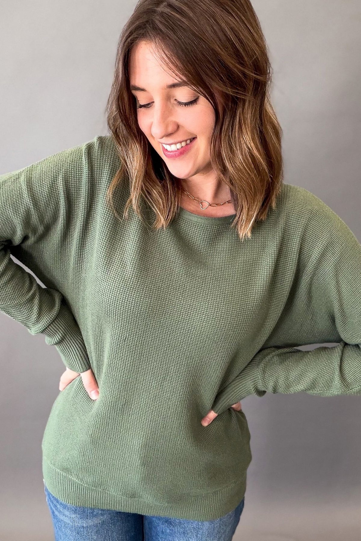 A woman wearing a sage green ribbed knit sweater with long sleeves and a crew neck, paired with blue jeans, posing against a neutral gray background.