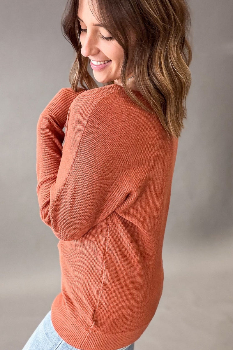 A woman wearing a textured, ribbed knit long-sleeve sweater in a warm terracotta or burnt orange color, shown from a side-back angle against a neutral gray studio background.