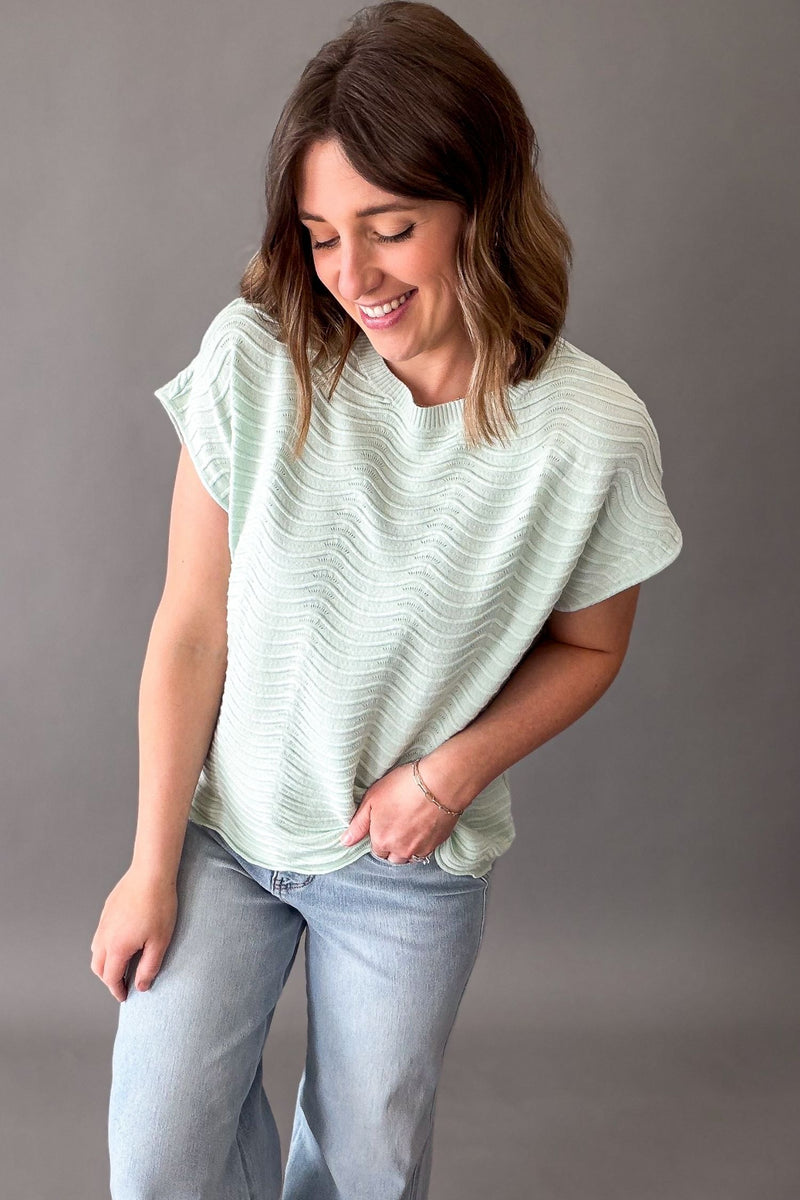 Woman wearing a light mint textured top and blue jeans against a gray background