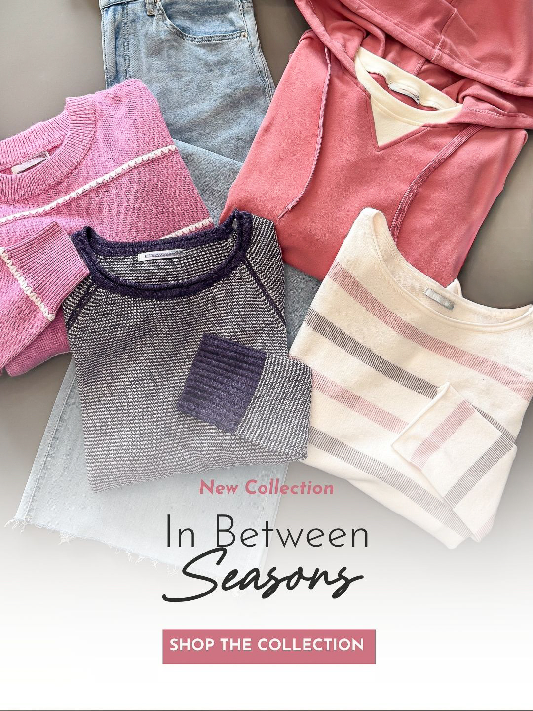 A styled flat lay of folded clothing on a neutral surface, featuring a pink knit sweater, light blue jeans, a coral pink hoodie, a gray striped long-sleeve top with dark purple trim, and a cream striped top. Text overlay reads 'New Collection In Between Seasons' with a pink 'SHOP THE COLLECTION' banner.