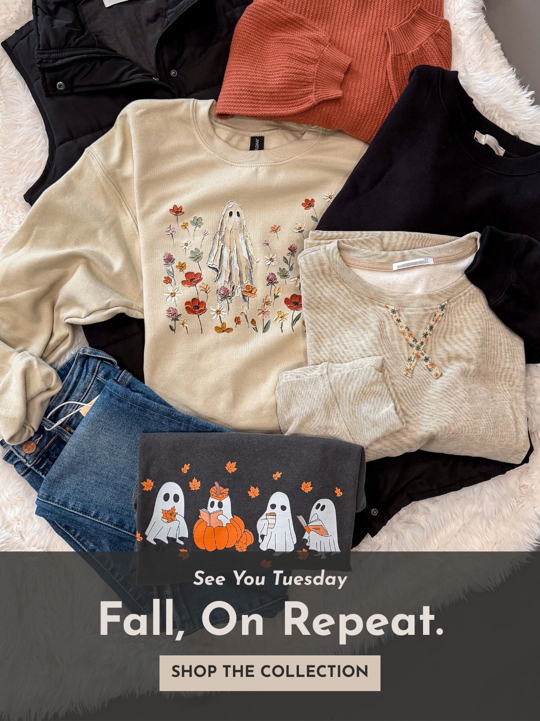 A styled flat lay of multiple fall clothing items on a white faux-fur surface: a beige crewneck sweatshirt with a ghost and floral print, a rust orange ribbed top, a black long-sleeve top, a cream knit top with a necklace, blue jeans, and a dark charcoal sweatshirt with a ghost graphic. Text at the bottom reads 'See You Tuesday Fall, On Repeat.