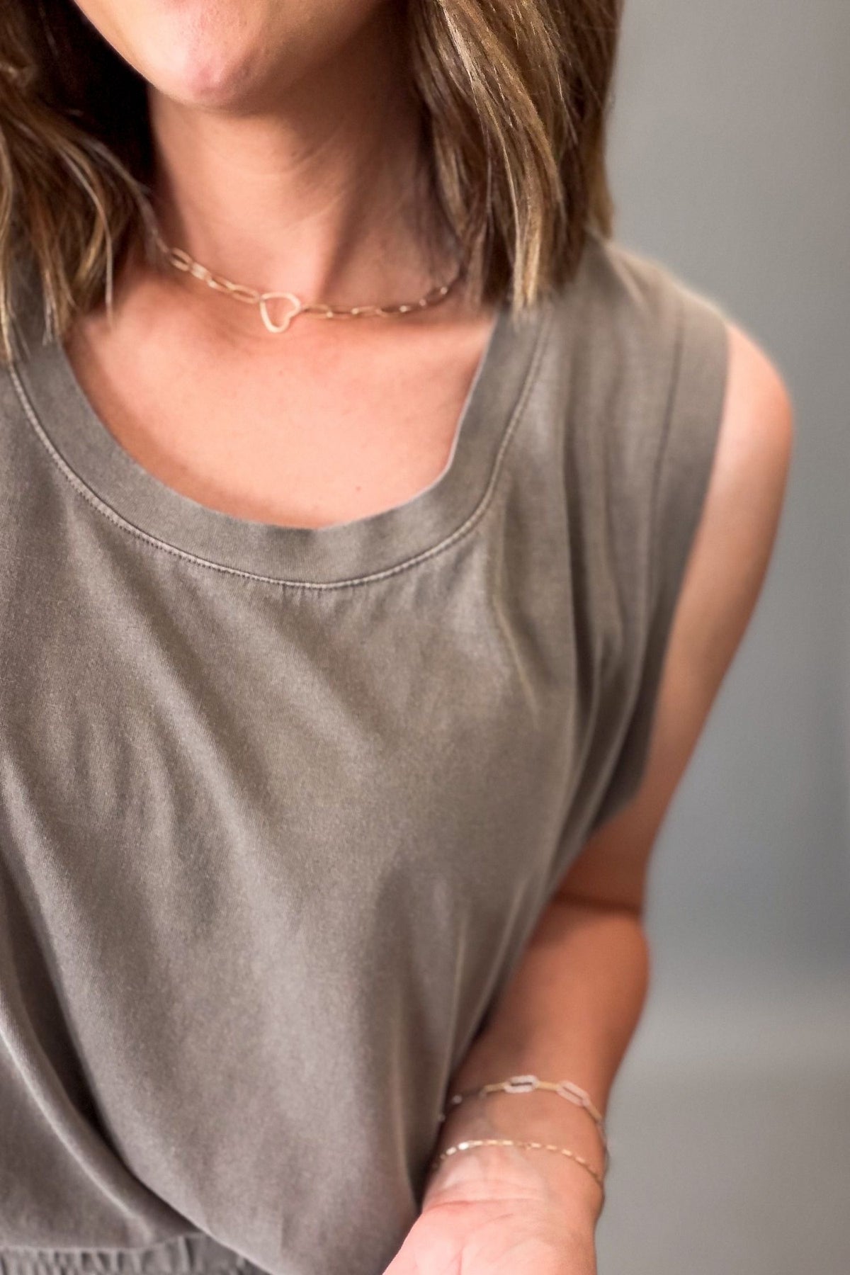 Close-up of a woman wearing a sleeveless mocha top and a delicate gold heart necklace, with her arm and hand visible against a soft gray background.