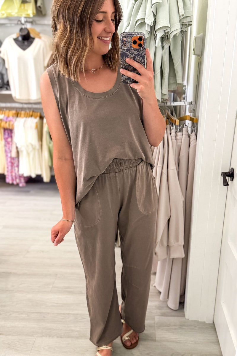 A woman takes a mirror selfie in a clothing boutique wearing a sleeveless mocha jumpsuit with a relaxed, wide-leg fit.