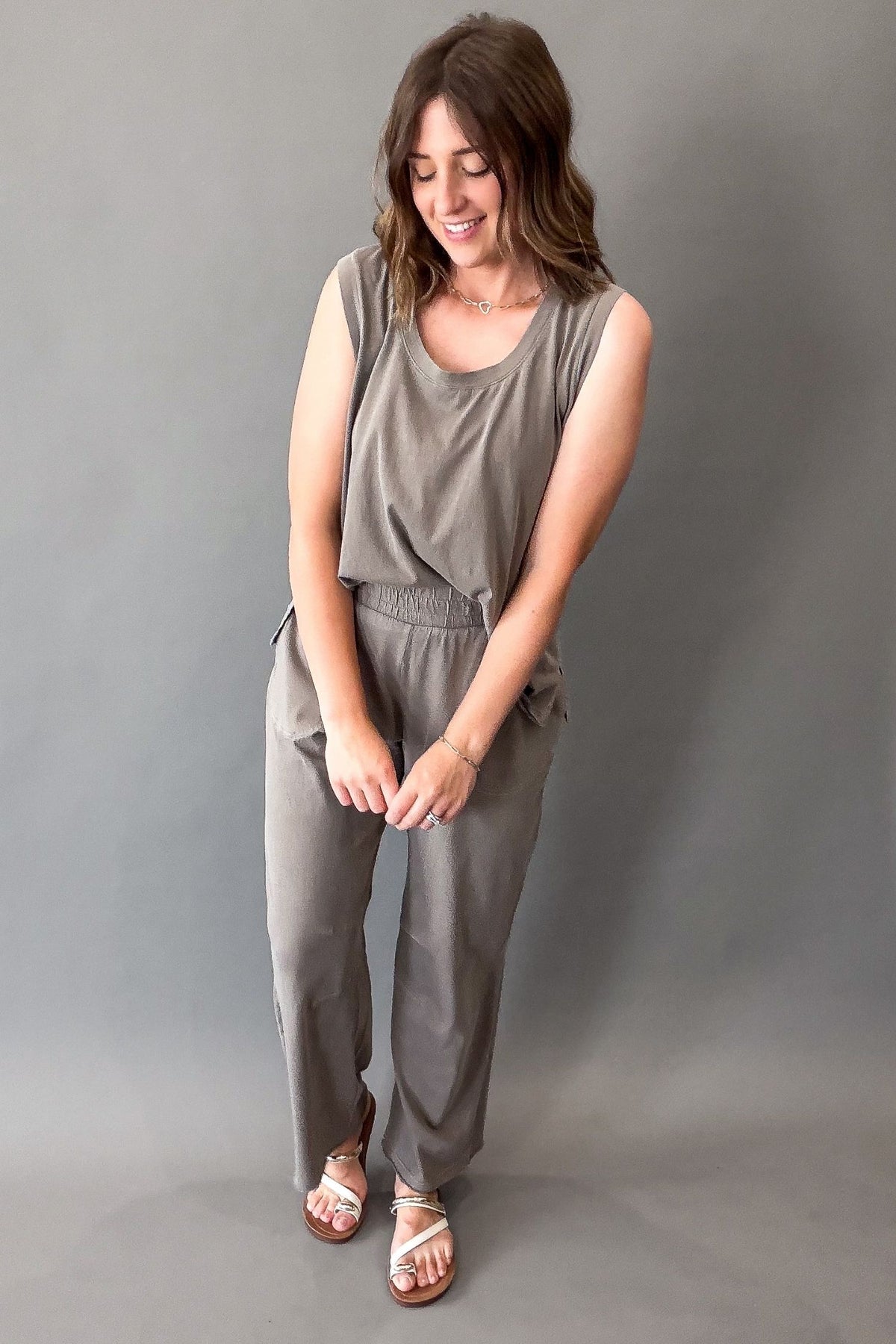 A woman modeling a sleeveless mocha jumpsuit with wide-leg pants and a matching fabric tie belt, paired with brown strappy sandals, against a solid gray studio backdrop.