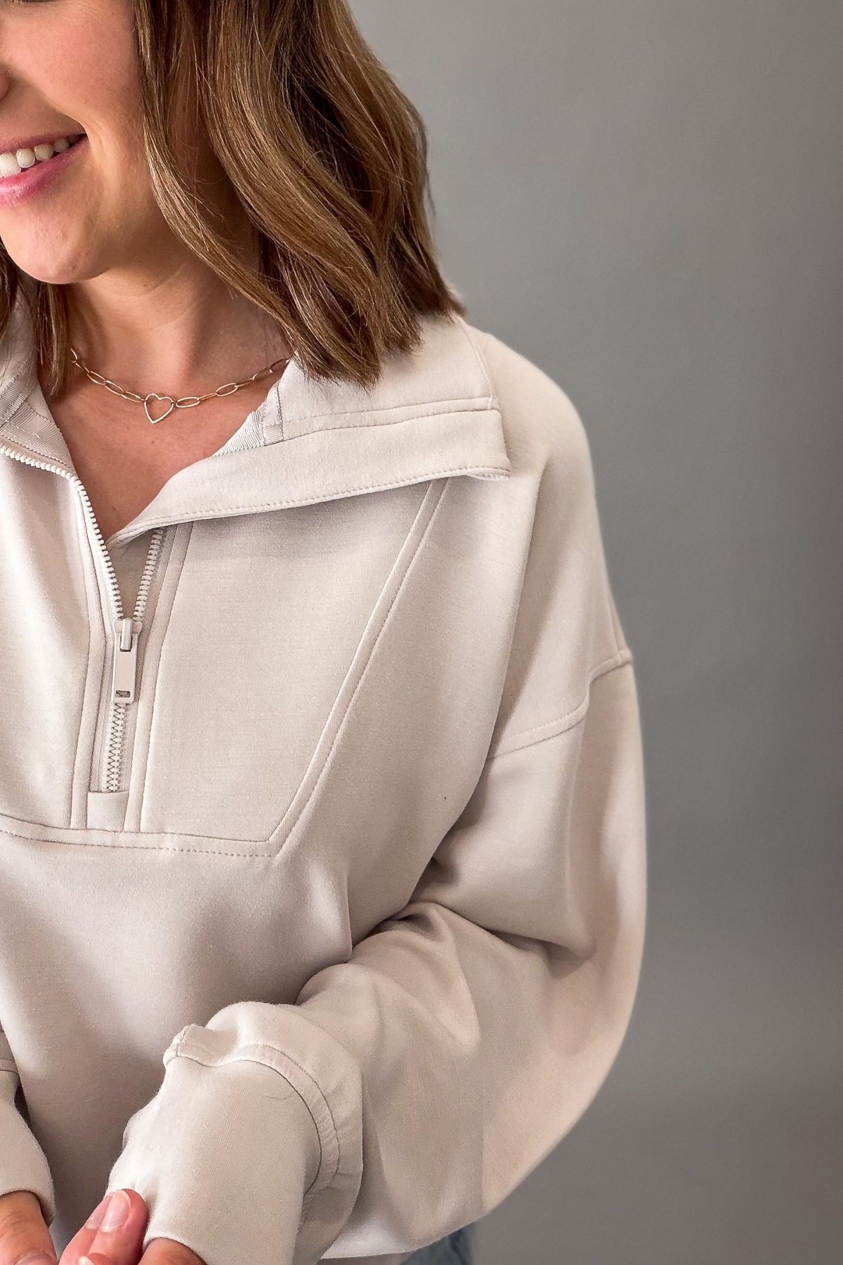 A close-up of a woman wearing a cream-colored half-zip sweatshirt with a wide collar, set against a solid gray background.