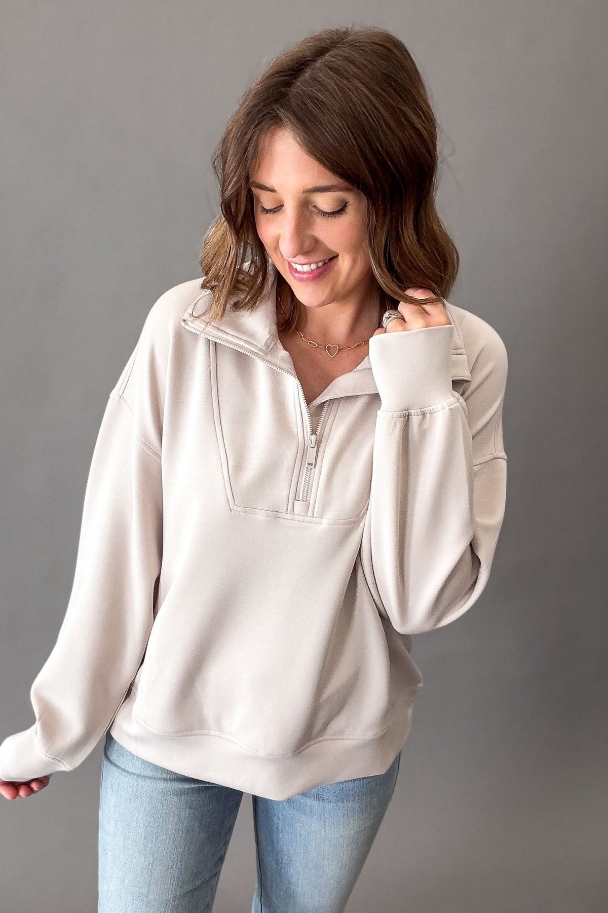 A woman wearing a mocha cream mock neck quarter-zip pullover sweatshirt and light blue jeans, standing against a solid gray studio background.