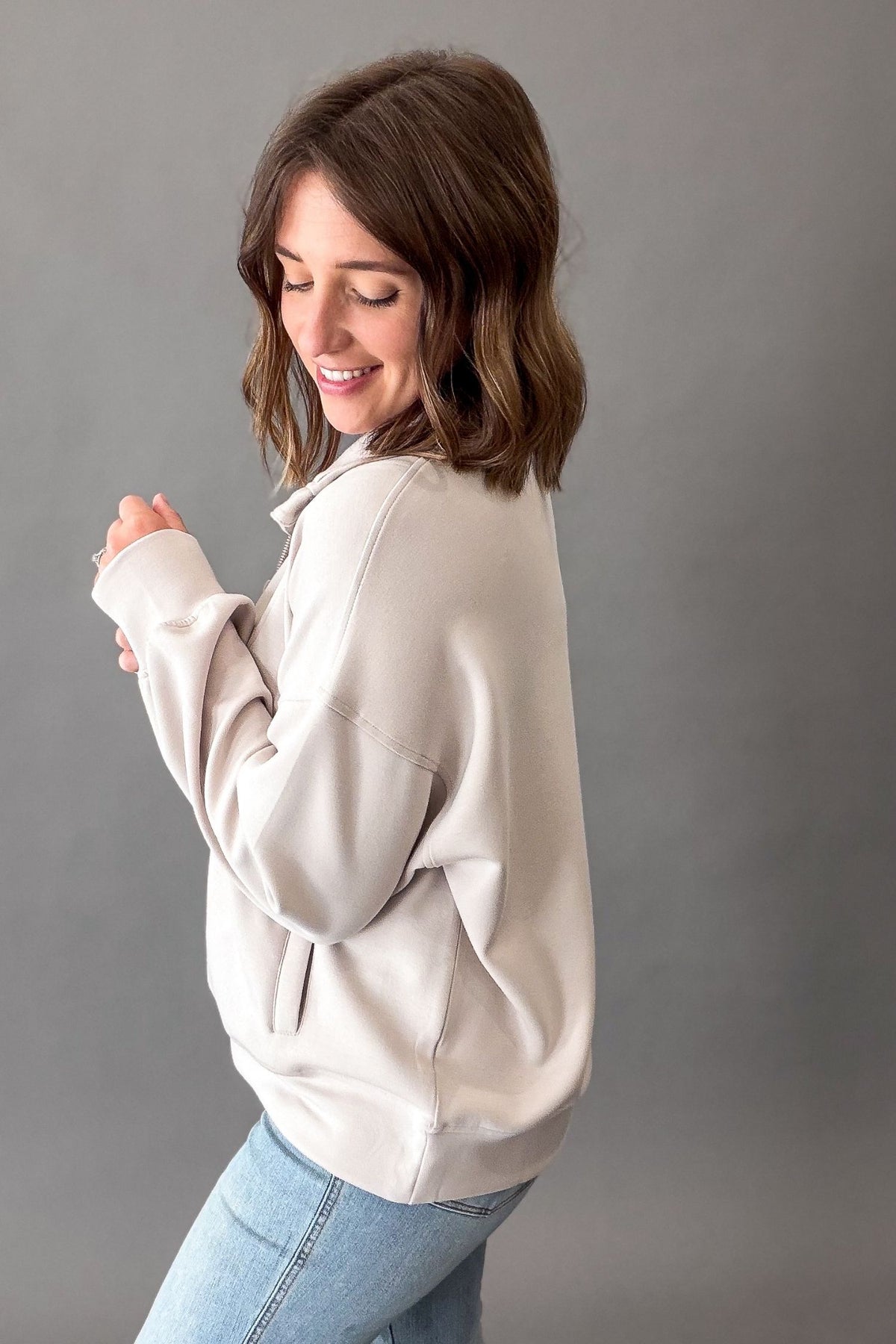 A woman with shoulder-length brown hair is shown from the side and back, wearing a mocha cream oversized quarter zip and light blue jeans against a solid gray studio background.