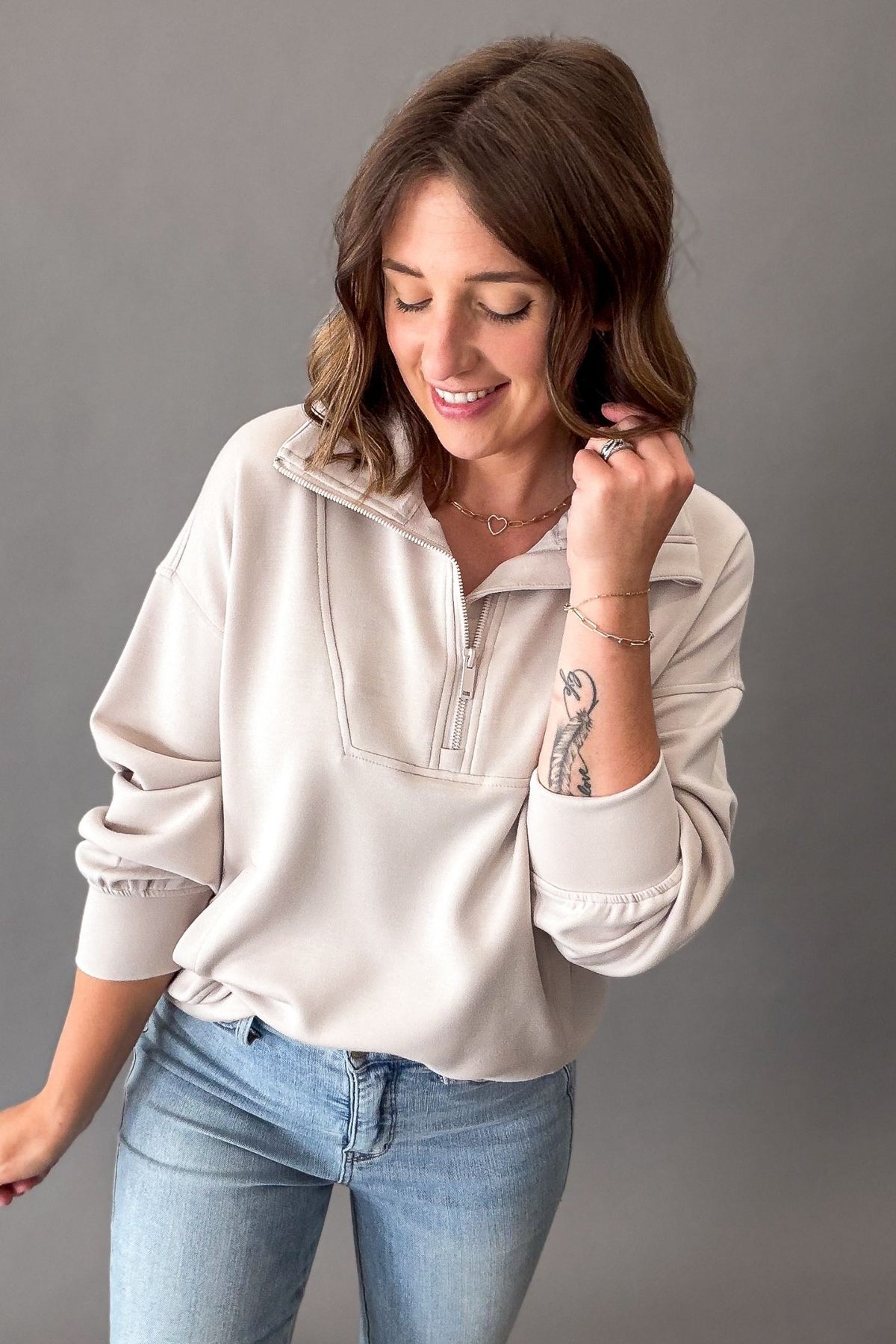 A woman wearing a mocha cream-colored quarter-zip sweatshirt and light-wash blue jeans, posing against a solid gray studio background.
