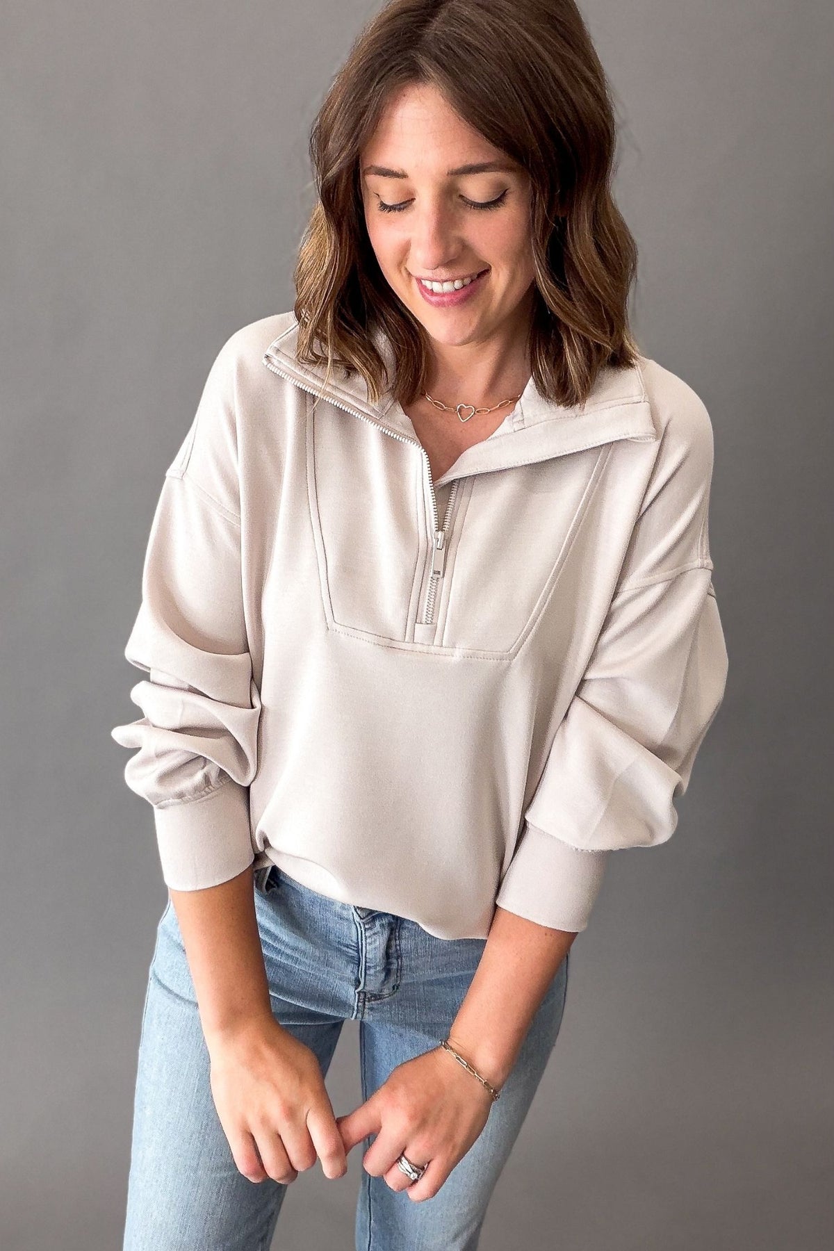 A woman wearing a mocha cream mock-neck pullover sweatshirt with a short zipper and gathered sleeves, paired with blue jeans, posing against a solid gray studio background.