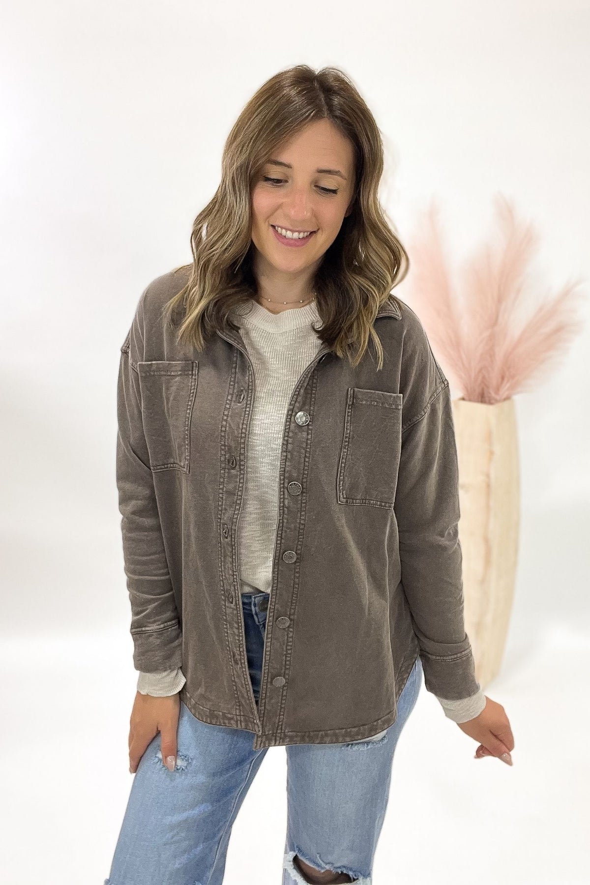 Woman wearing a brown jacket over a white shirt and blue jeans, standing against a white background with a pink decorative item.