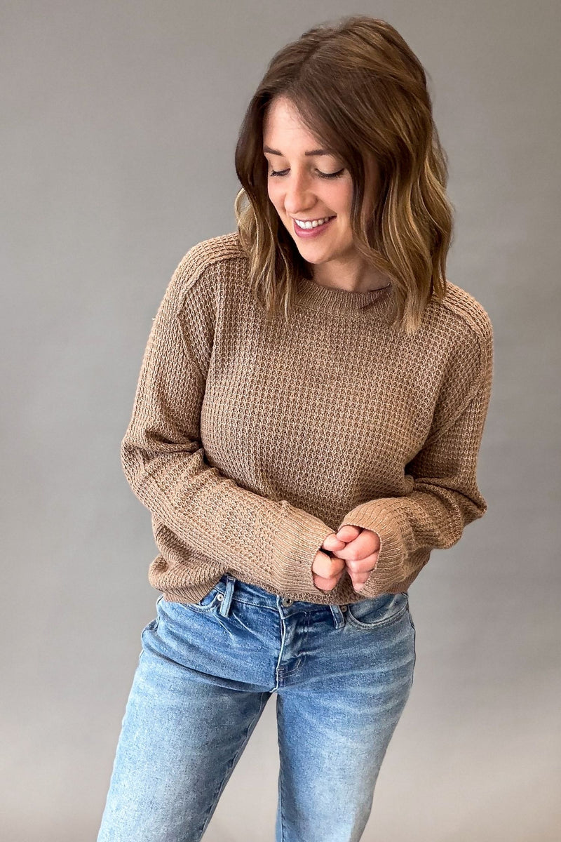 A woman wearing a mocha waffle-knit sweater and blue jeans, standing against a neutral gray studio background.