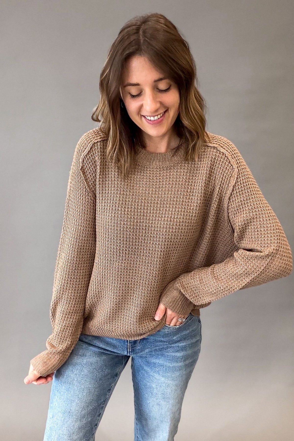 A woman wearing a mocha waffle-knit long-sleeve sweater and blue jeans, standing against a plain gray studio background.