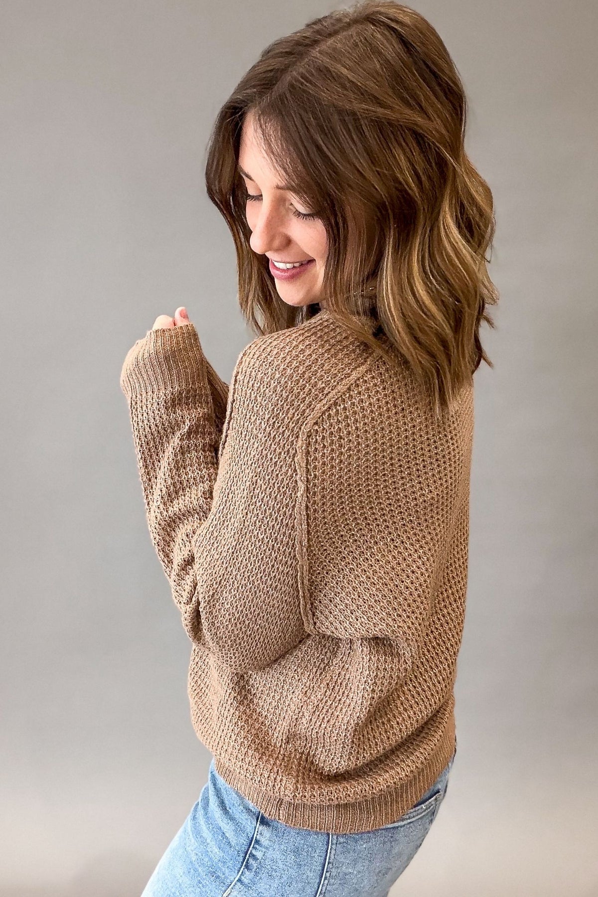 A woman wearing a textured, waffle-knit mocha sweater and light blue jeans, photographed from a side-back angle against a neutral gray studio background.