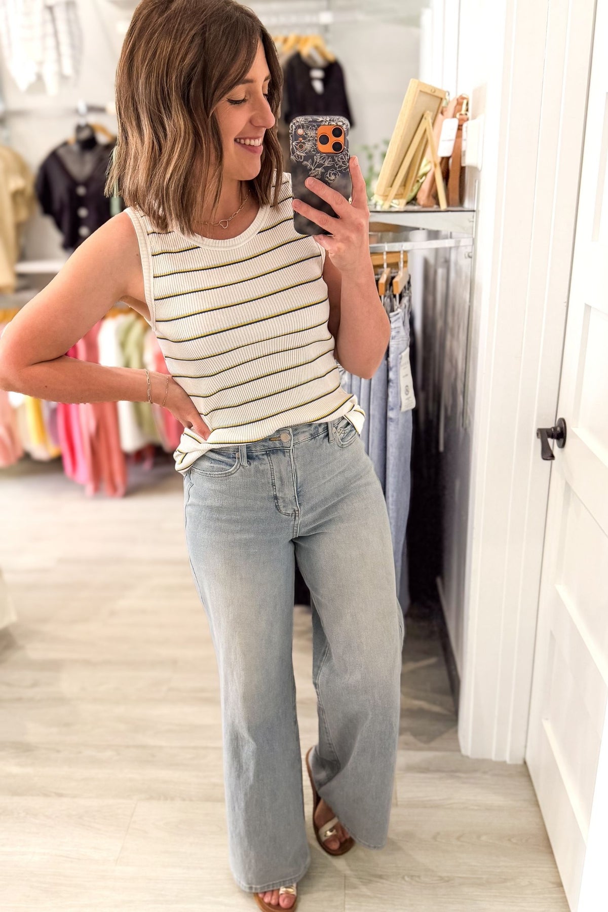 Woman taking a mirror selfie wearing a striped tank top and light blue jeans in a clothing store.