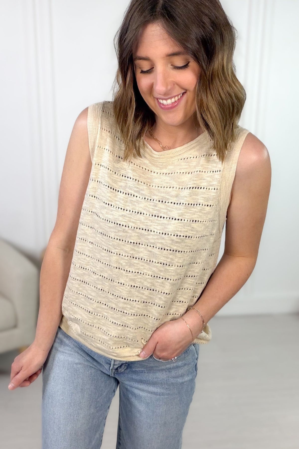 Woman wearing a beige sleeveless top with a subtle pattern, standing against a white wall.