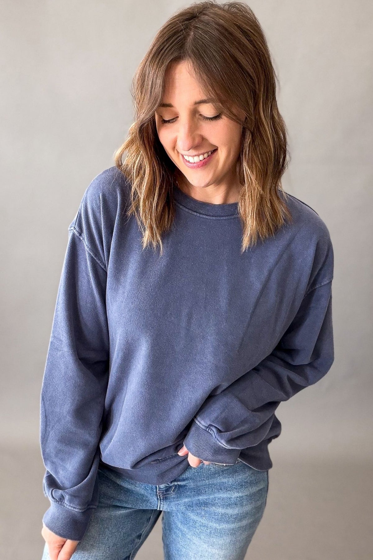 A woman with shoulder-length brown hair smiling and looking down, wearing a washed slate blue crewneck sweatshirt and blue jeans against a neutral gray studio background.