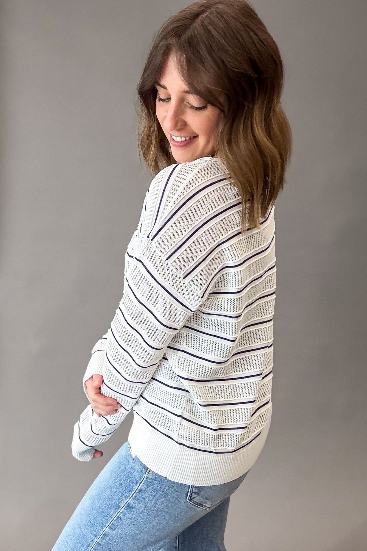 Woman wearing a white textured pullover with navy stripes and blue jeans against a gray background