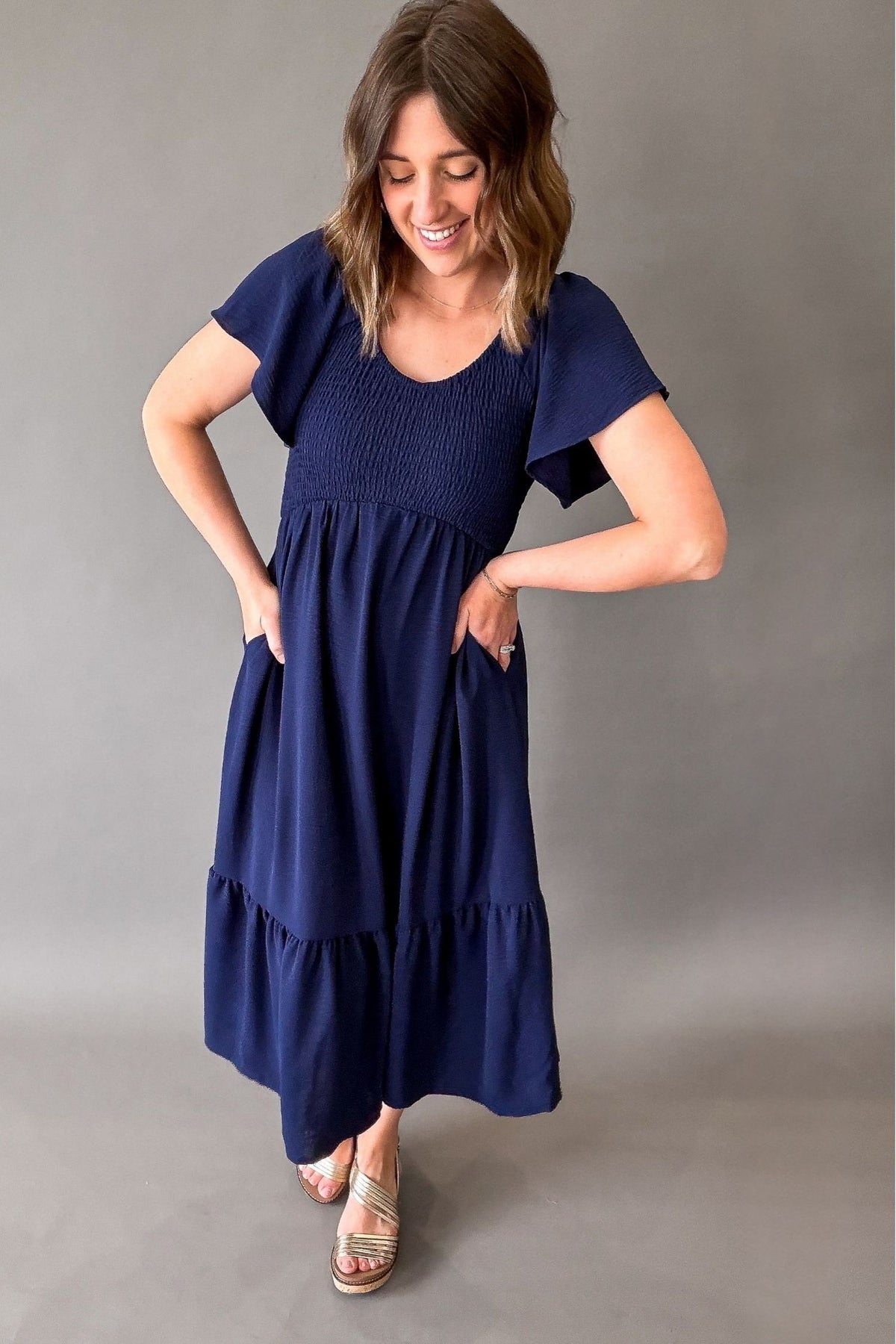 Woman wearing a navy blue dress with a smocked bust area and flutter sleeves, against a gray background showing pockets.