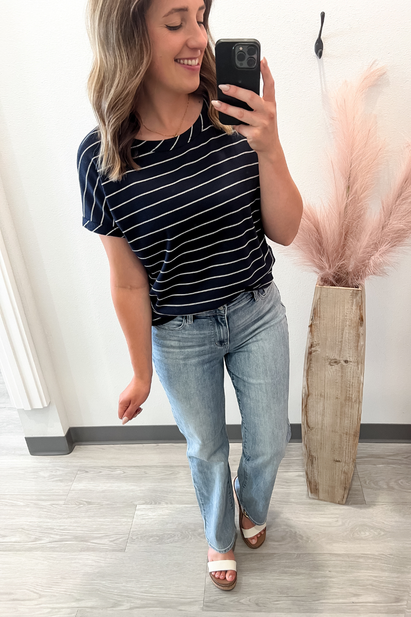 Woman taking a mirror selfie wearing a striped shirt and jeans, standing in a room with a plant in the background.