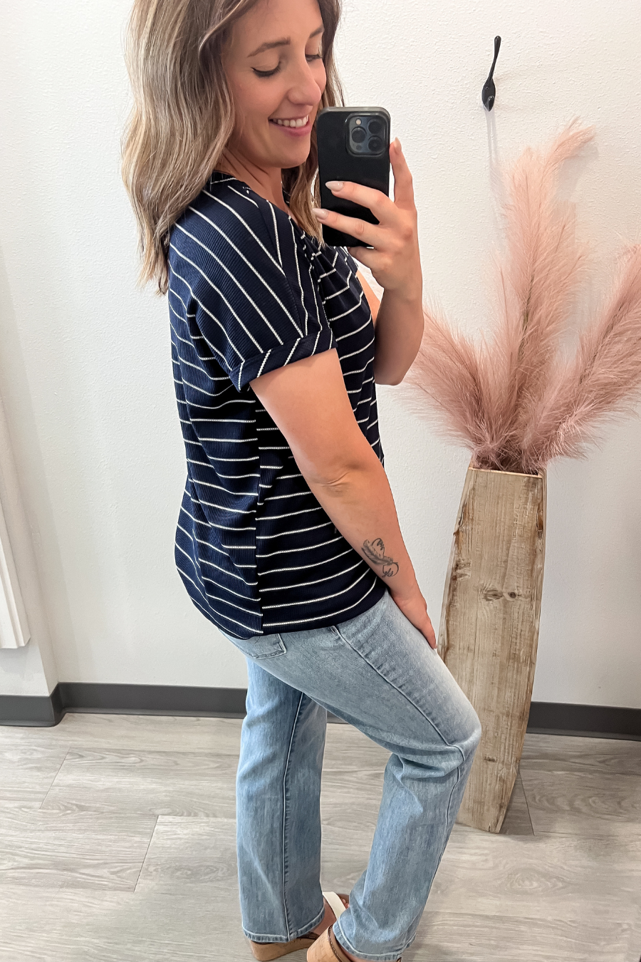 Woman taking a mirror selfie wearing a striped shirt and jeans, standing next to a decorative plant.