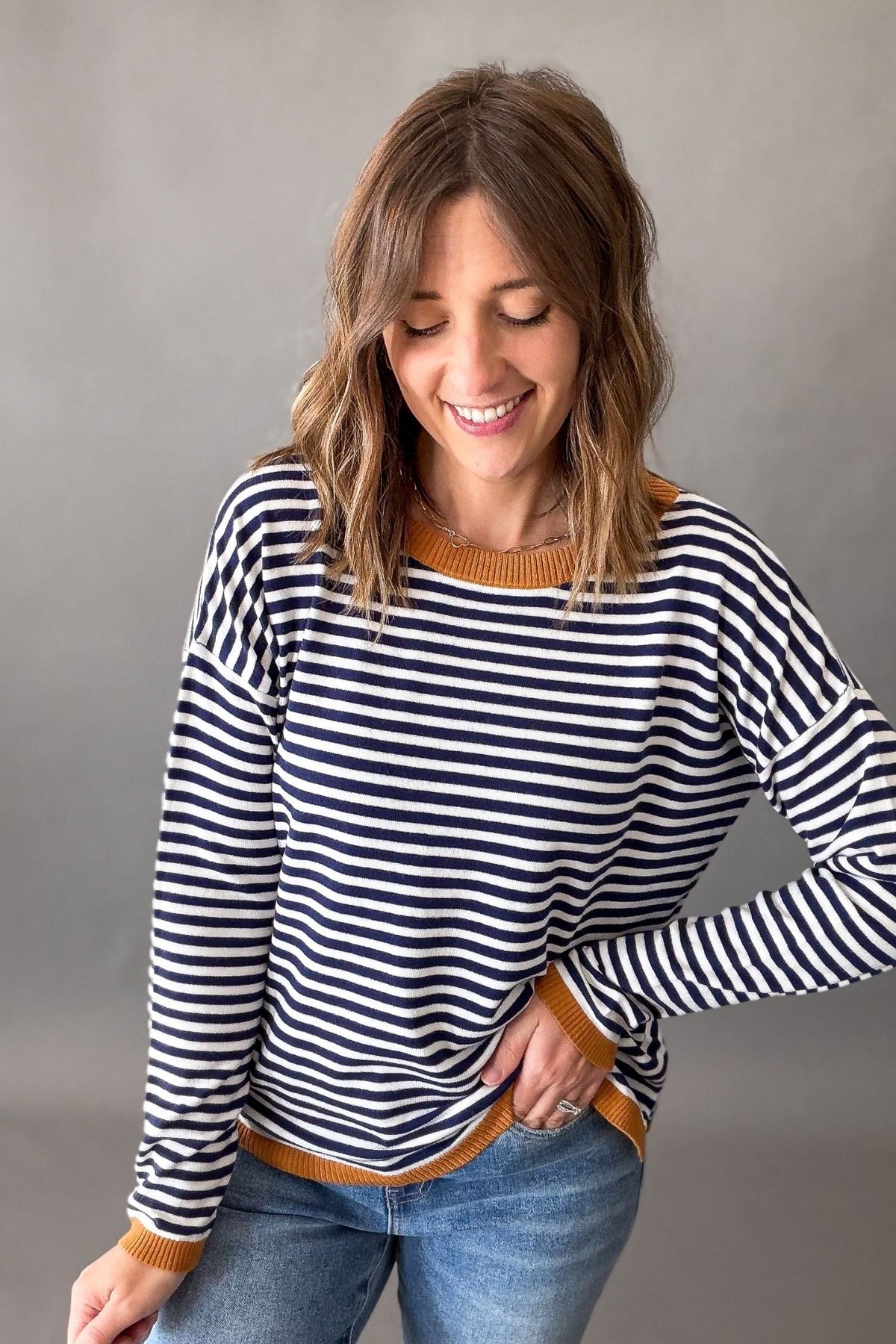 A woman wearing a navy and white horizontally striped long-sleeve sweater with a mustard yellow ribbed crew neck, cuffs, and hem, paired with blue jeans, against a neutral gray studio background.
